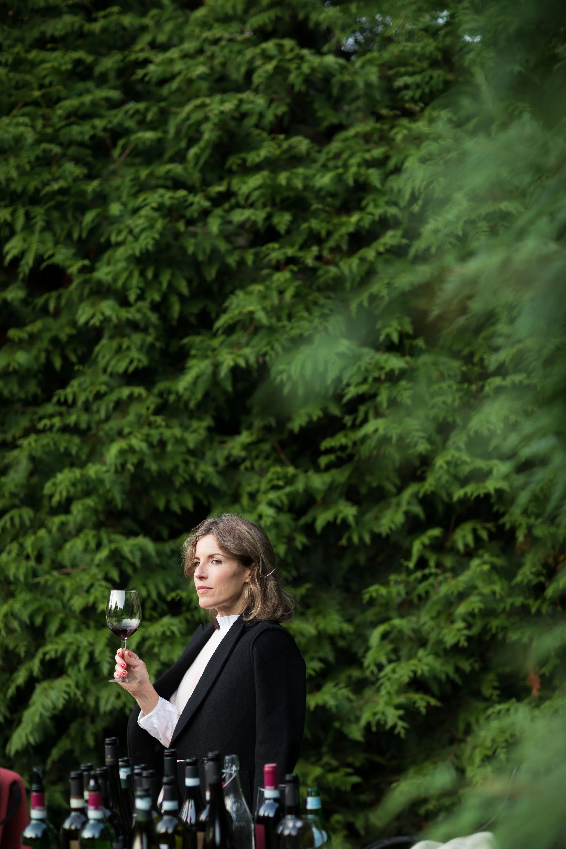 A woman holding a glass of red wine stands outdoors with a large green leafy shrub in the background. There are several wine bottles on a table in front of her.
