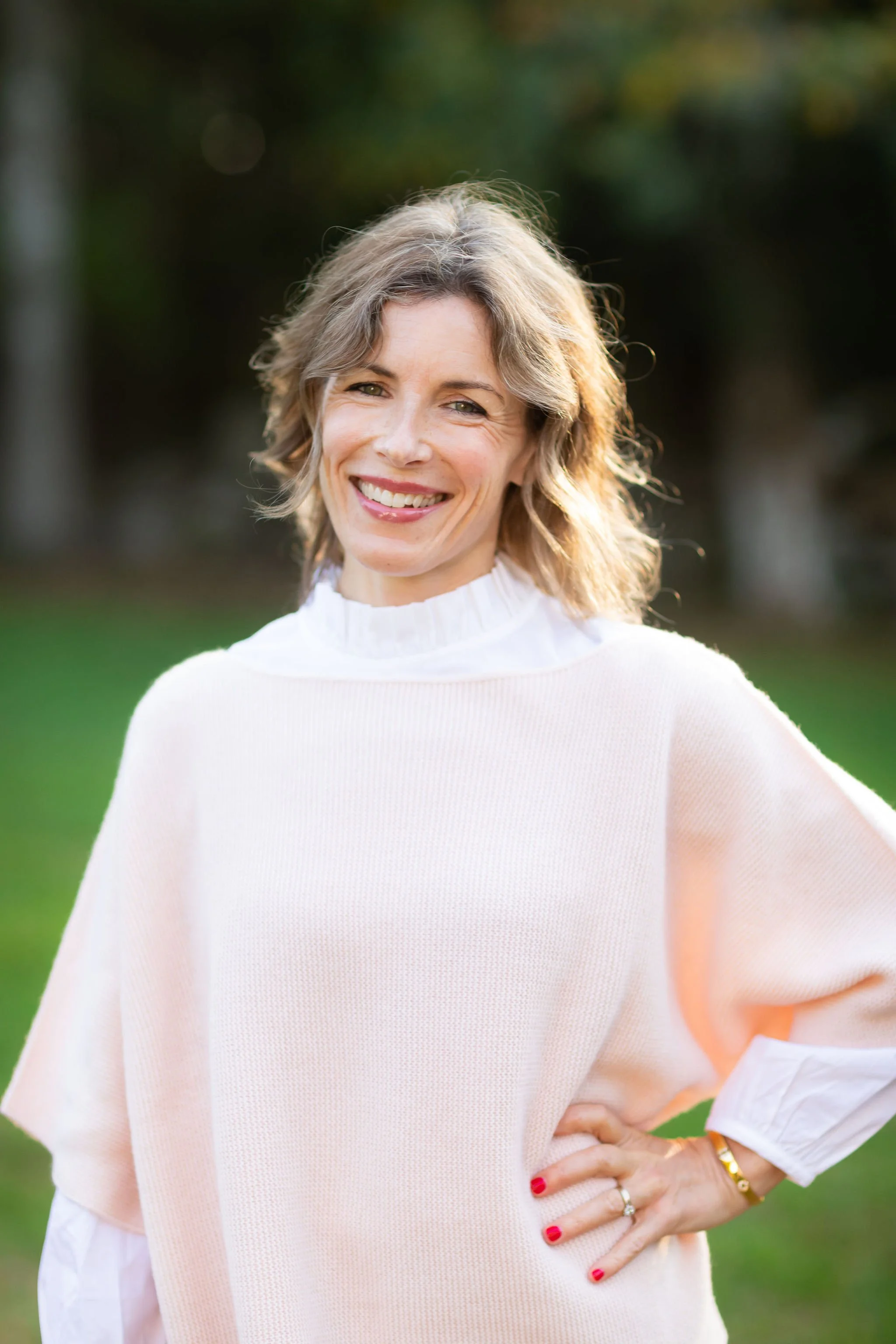 A smiling woman with shoulder-length blonde hair, wearing a white turtleneck sweater and a light pink poncho, standing outdoors in a park with greenery in the background.