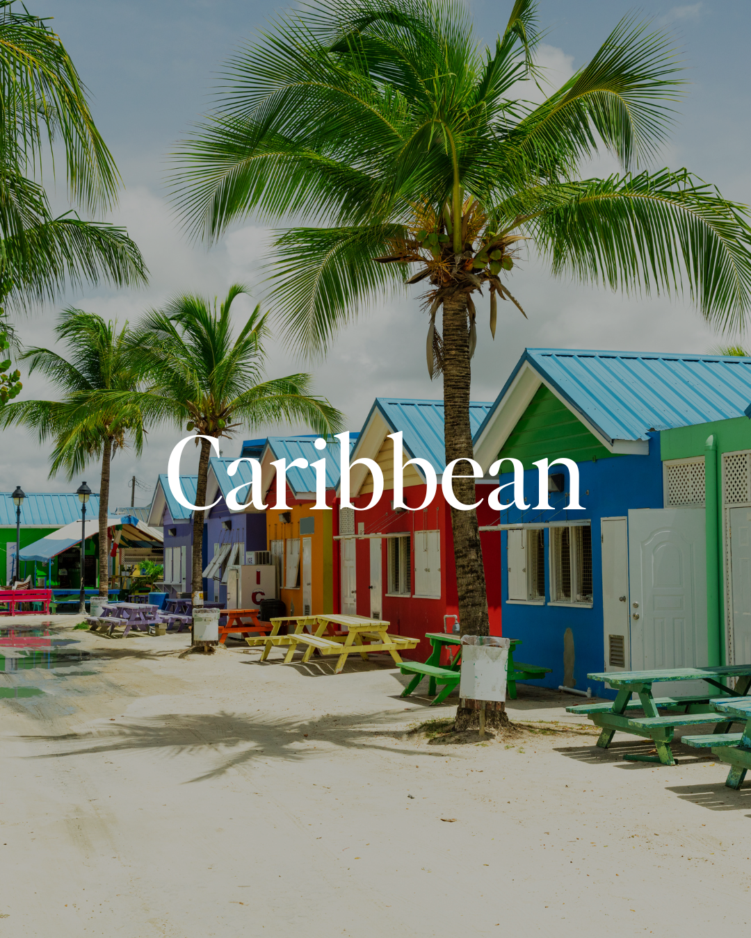 Colorful beachside buildings with blue, green, red, and purple walls, surrounded by palm trees, with sand and outdoor picnic tables in the foreground in the Caribbean.