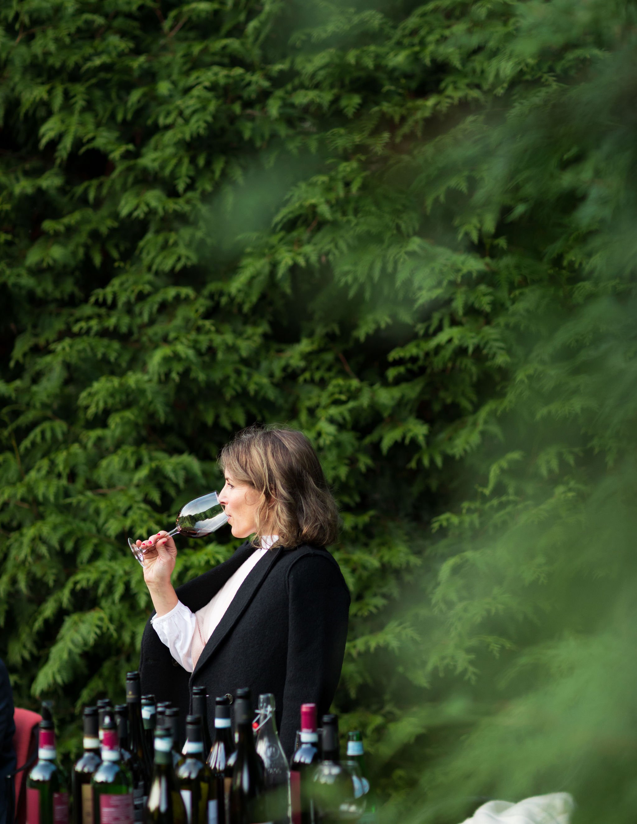 Woman tasting red wine outdoors with green foliage in background, surrounded by wine bottles on a table.