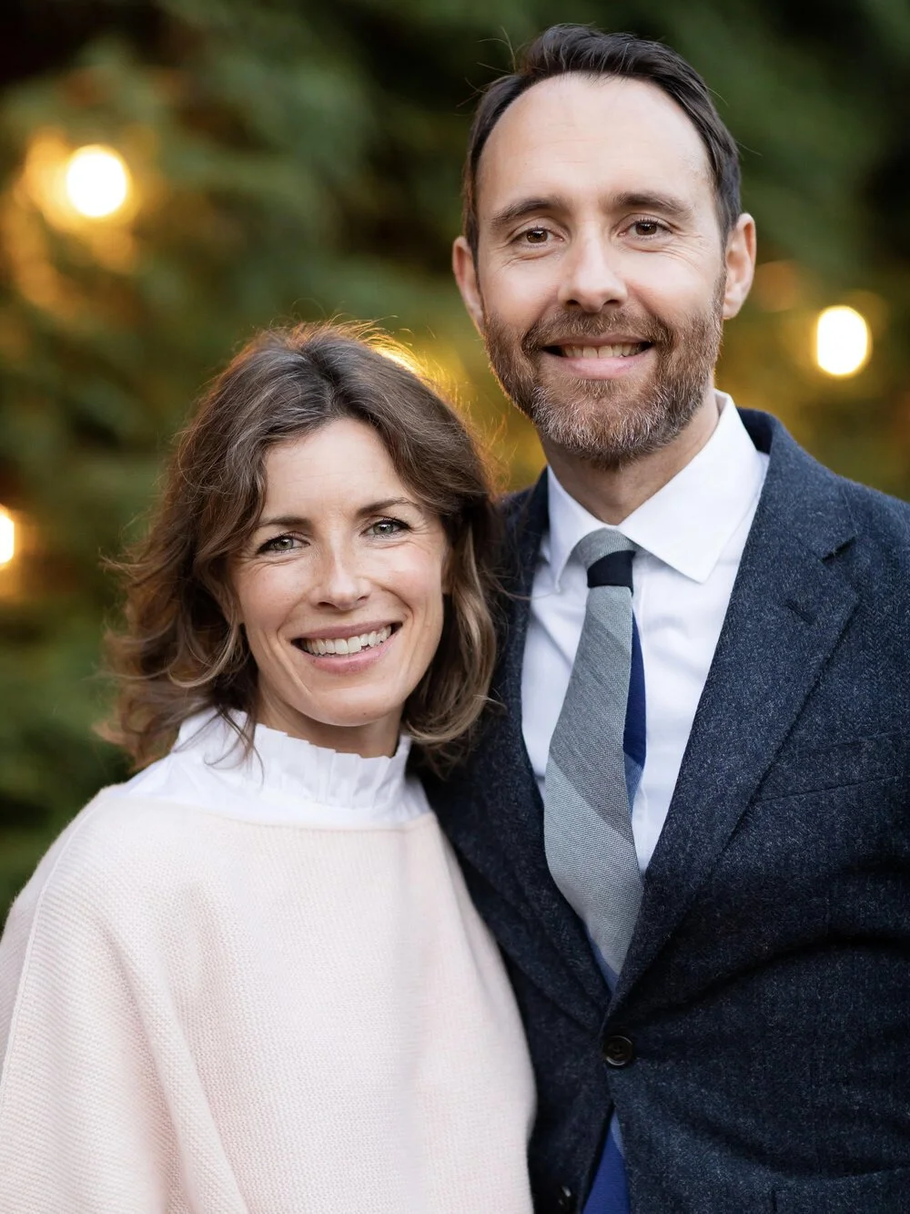 A smiling couple posing outdoors with blurred holiday lights and greenery in the background.