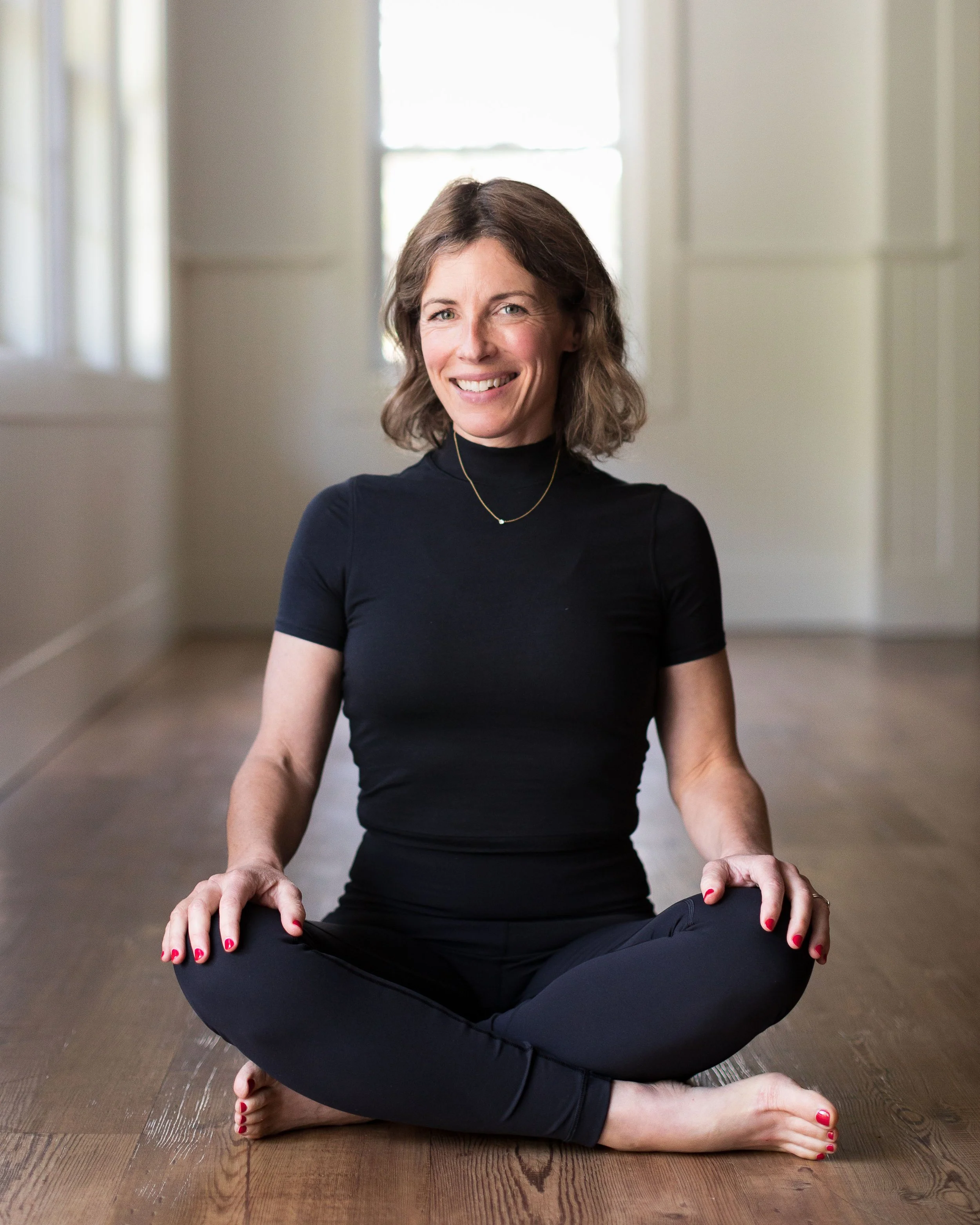 A woman practicing yoga or meditation, sitting cross-legged on a wooden floor in a bright room with large windows, wearing a black turtleneck and black leggings.