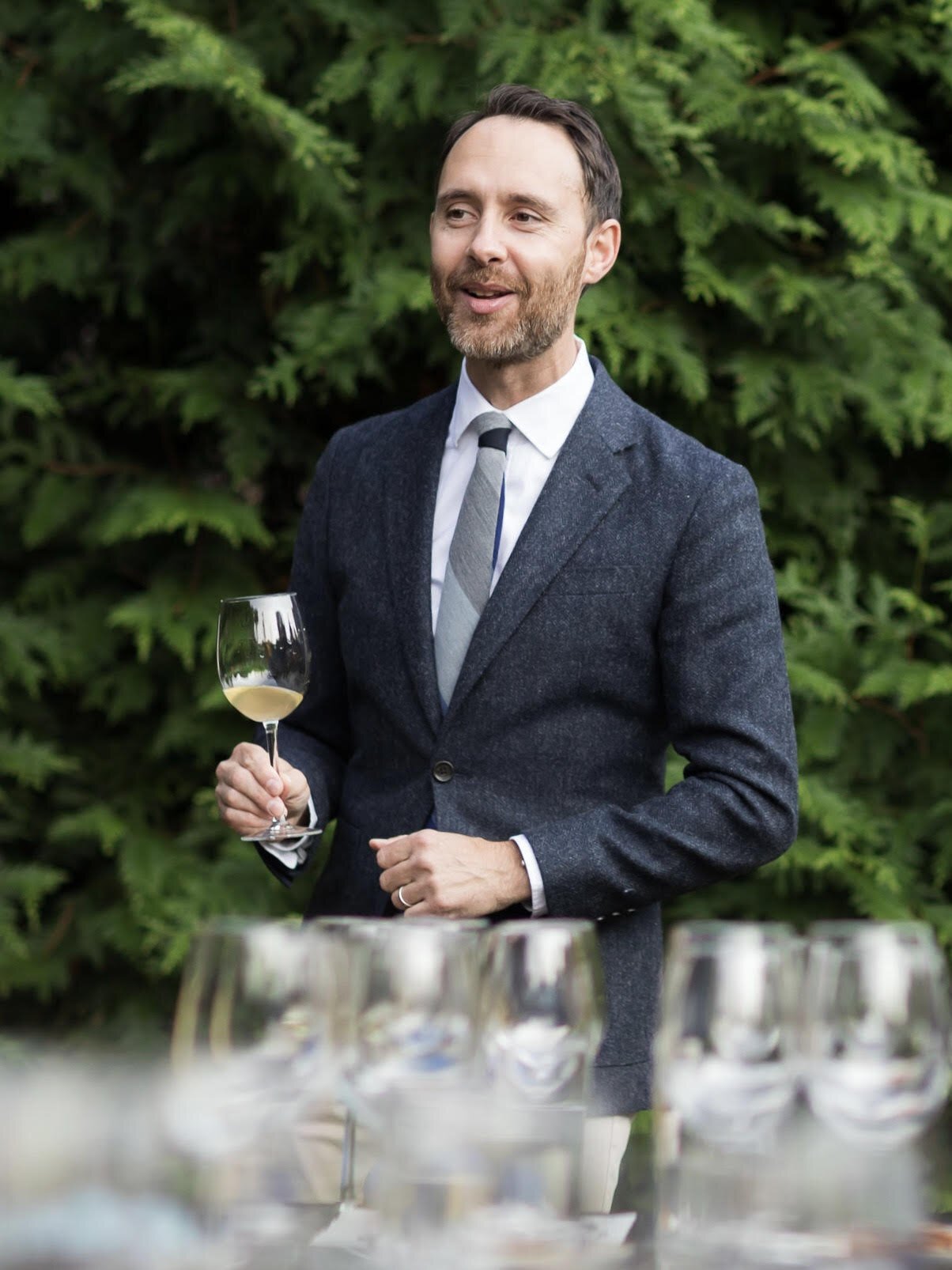 A man in a dark gray suit holding a glass of white wine at an outdoor gathering with a green leafy background and several empty glasses on the table.