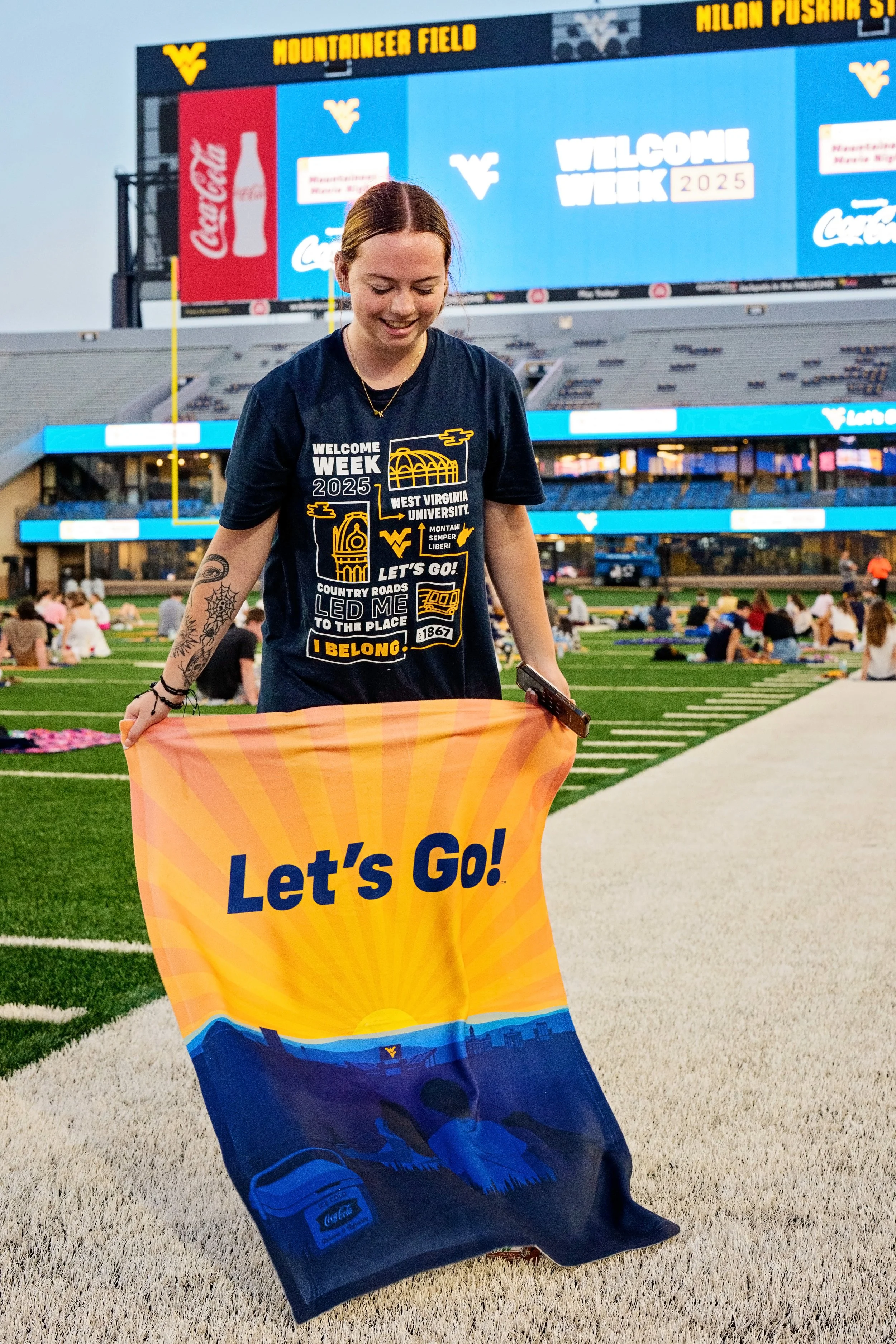 A girl wearing the 2025 tshirt on the football field holding up the WVU flag