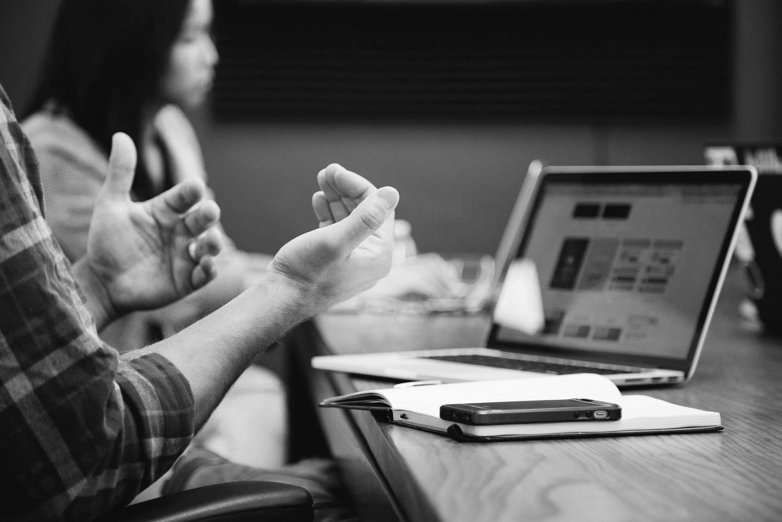 A meeting with a person explaining while sitting at a table with a laptop, notebook, and smartphone.