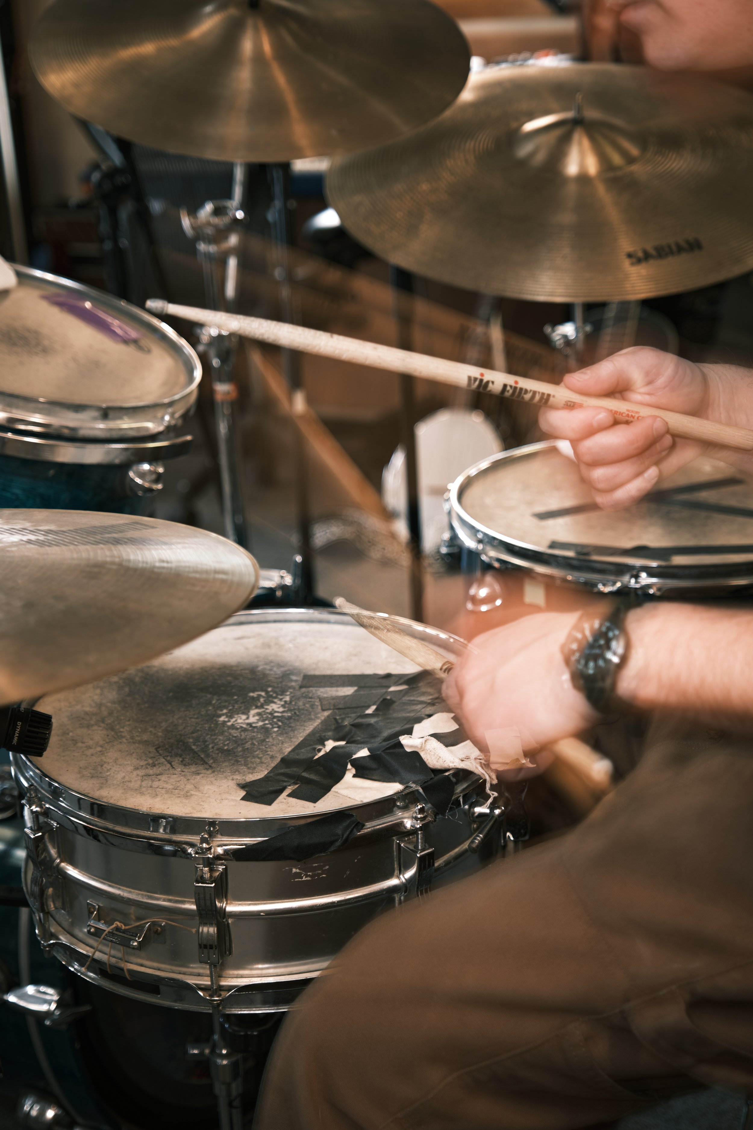 Close-up of a person playing drums, holding drumsticks near the snare drum with worn drum heads and cymbals in the background.