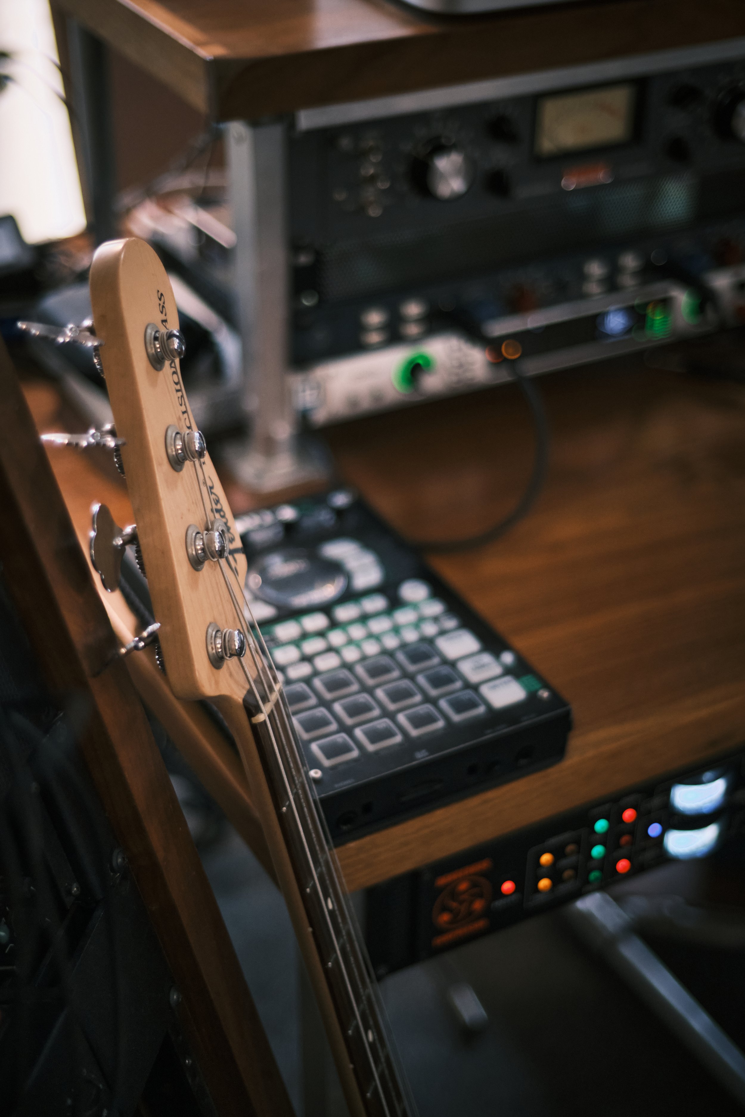 Close-up of a wooden guitar headstock with tuning pegs, resting against a desk with music production equipment including a MIDI controller, audio interface, and mixer.