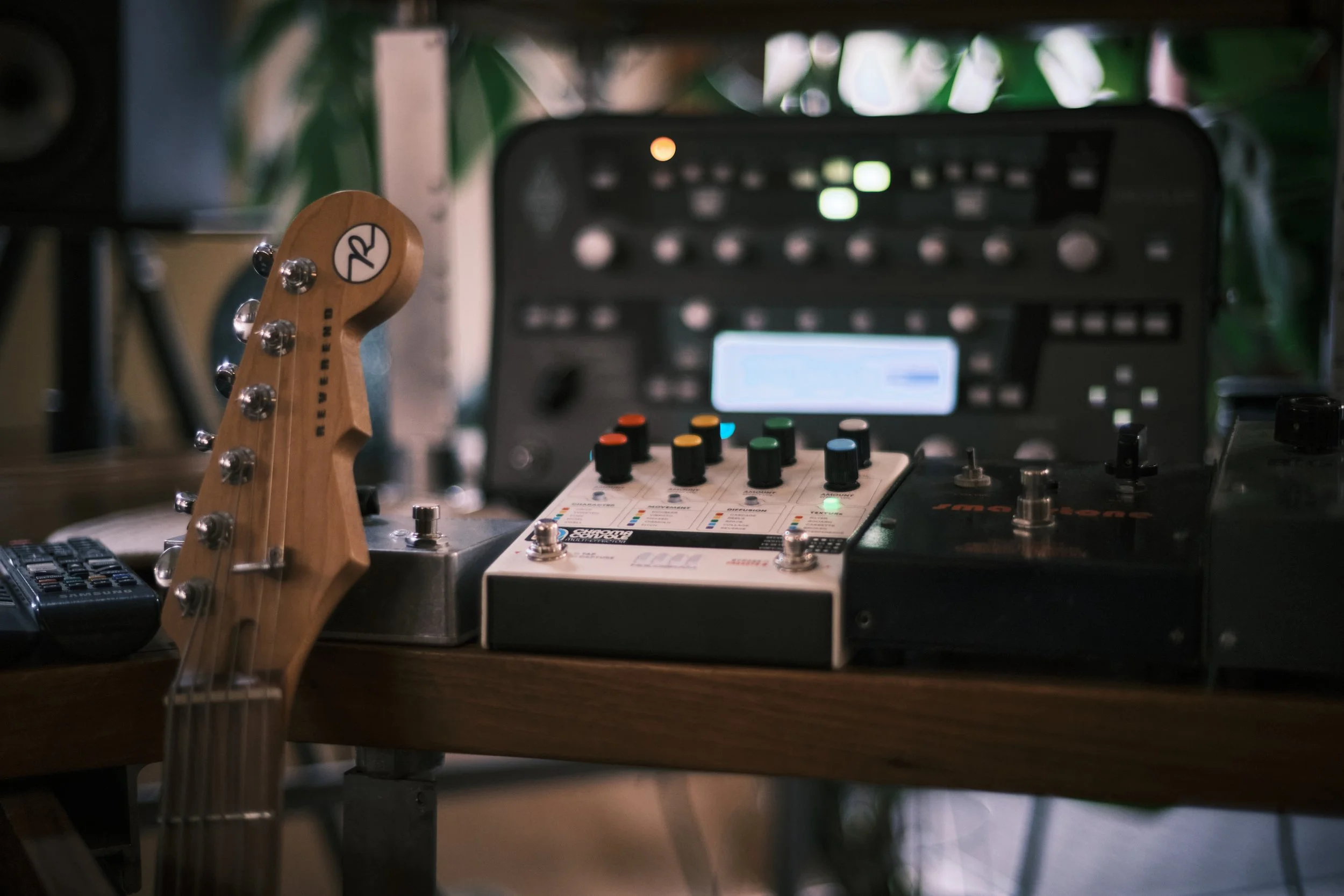 Guitar headstock, audio mixer, and effects pedals on a wooden table in a music studio.