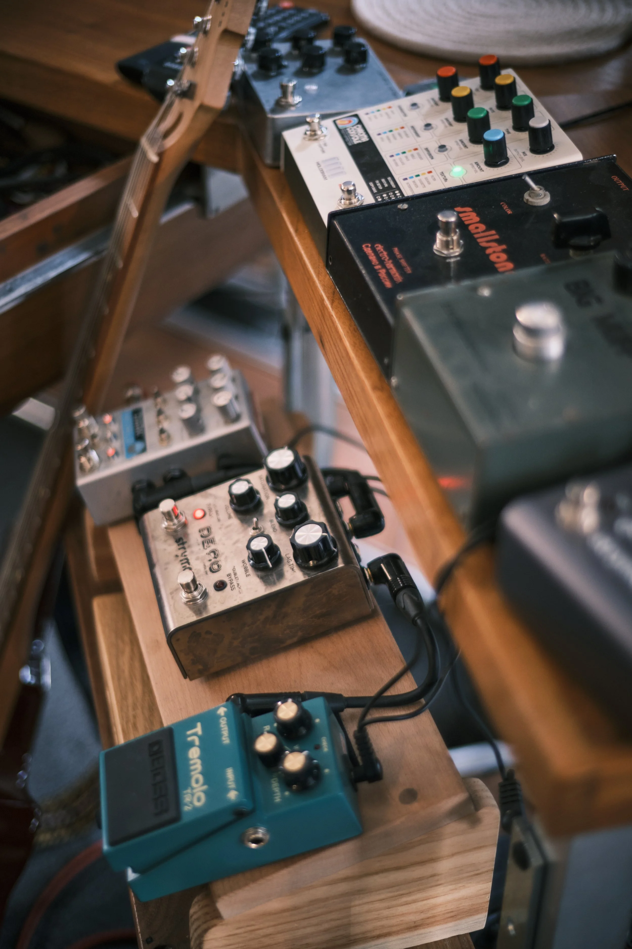 Various guitar effect pedals arranged on a wooden table, including a T Rex Tonebug, Big Ear, B'yoo, and other pedals, with a guitar neck visible on the left.