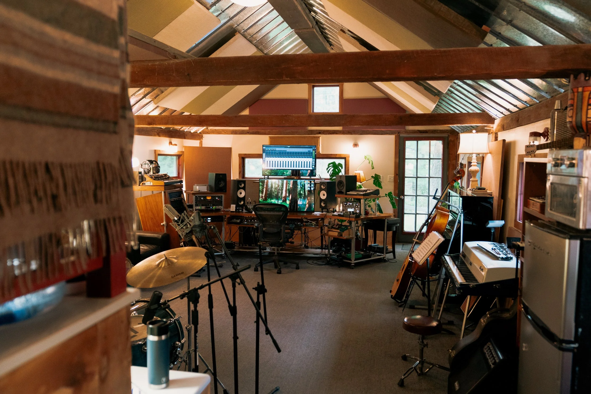 Music recording studio with multiple musical instruments including drums, a keyboard, guitars, and sheets of music, and audio equipment such as speakers and a computer monitor, inside a room with exposed wooden beams and large windows.