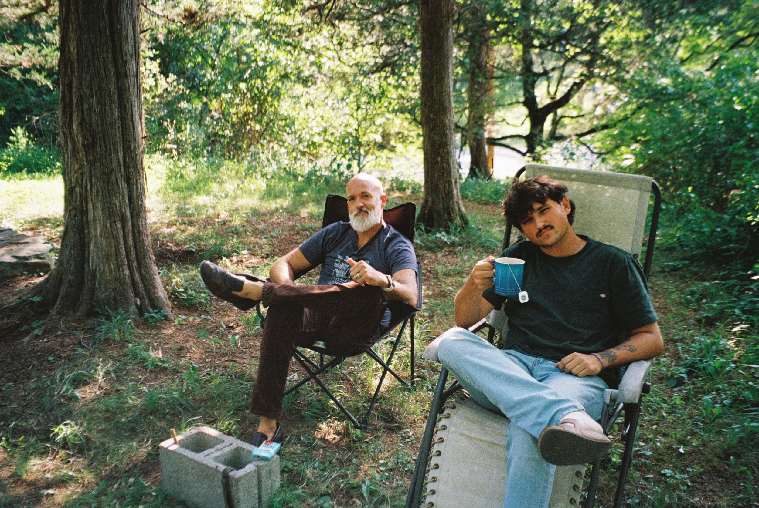 Two men sitting in camping chairs in a wooded area, one holding a blue mug, surrounded by trees and greenery.