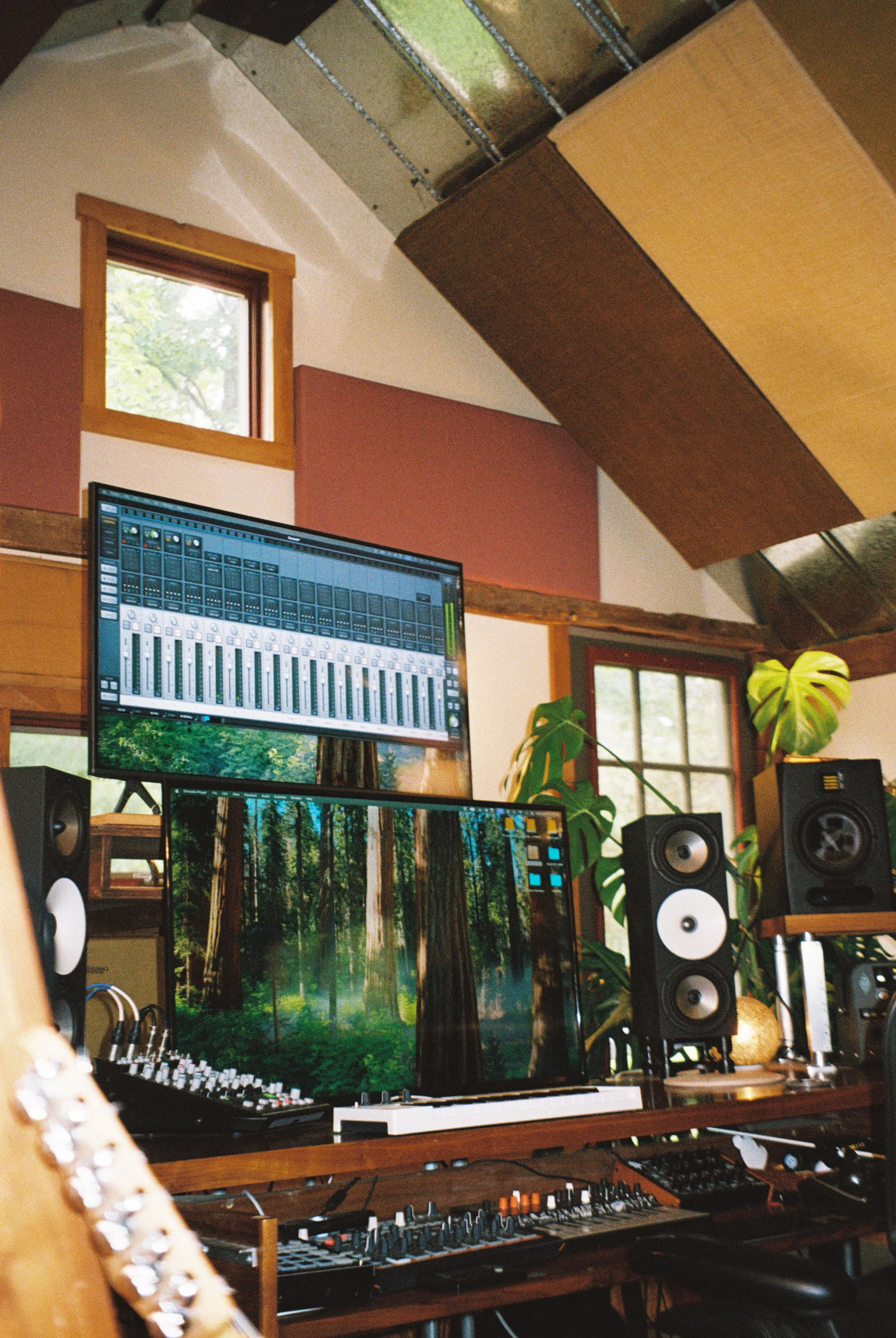Inside a music studio with computer monitors displaying a forest background, speakers, audio equipment, and soundproofing panels.