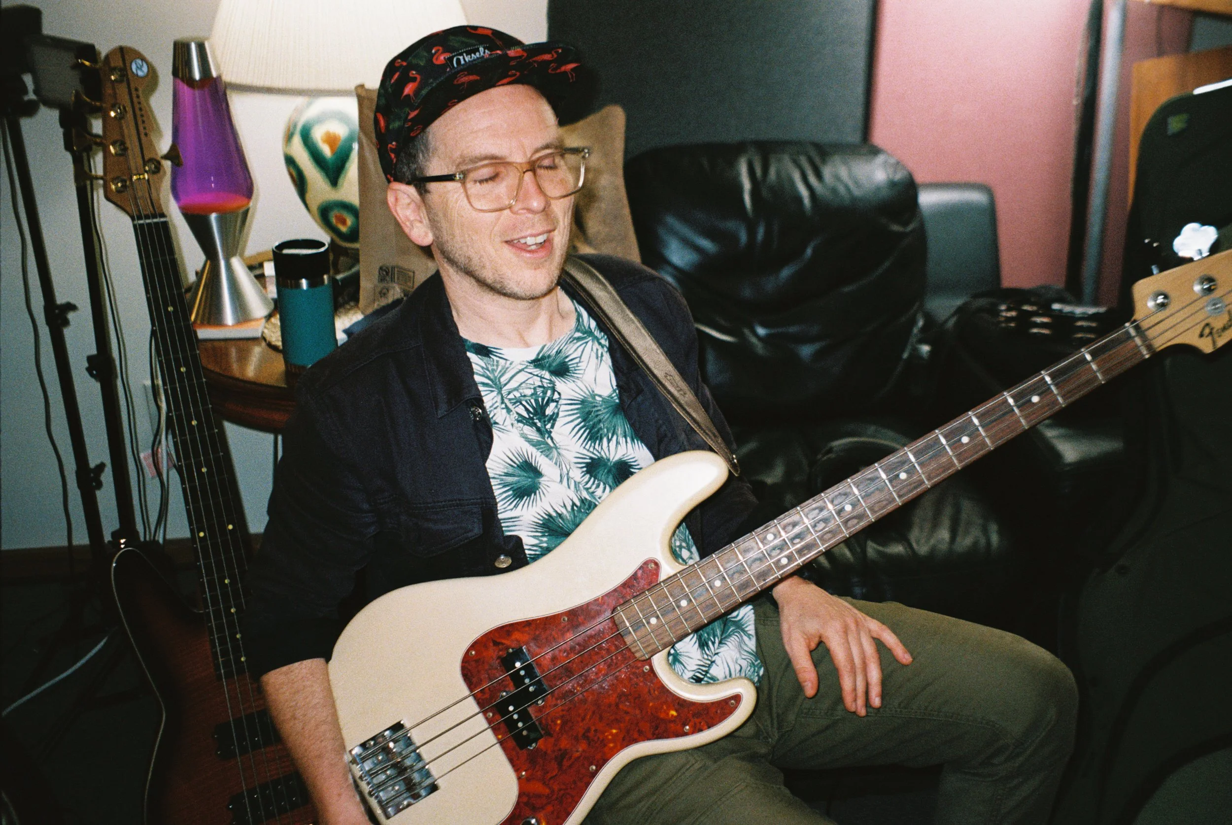 A man playing an electric bass guitar in a cozy room, wearing glasses, a patterned shirt, a jacket, and a cap, with musical equipment and colorful decor around him.