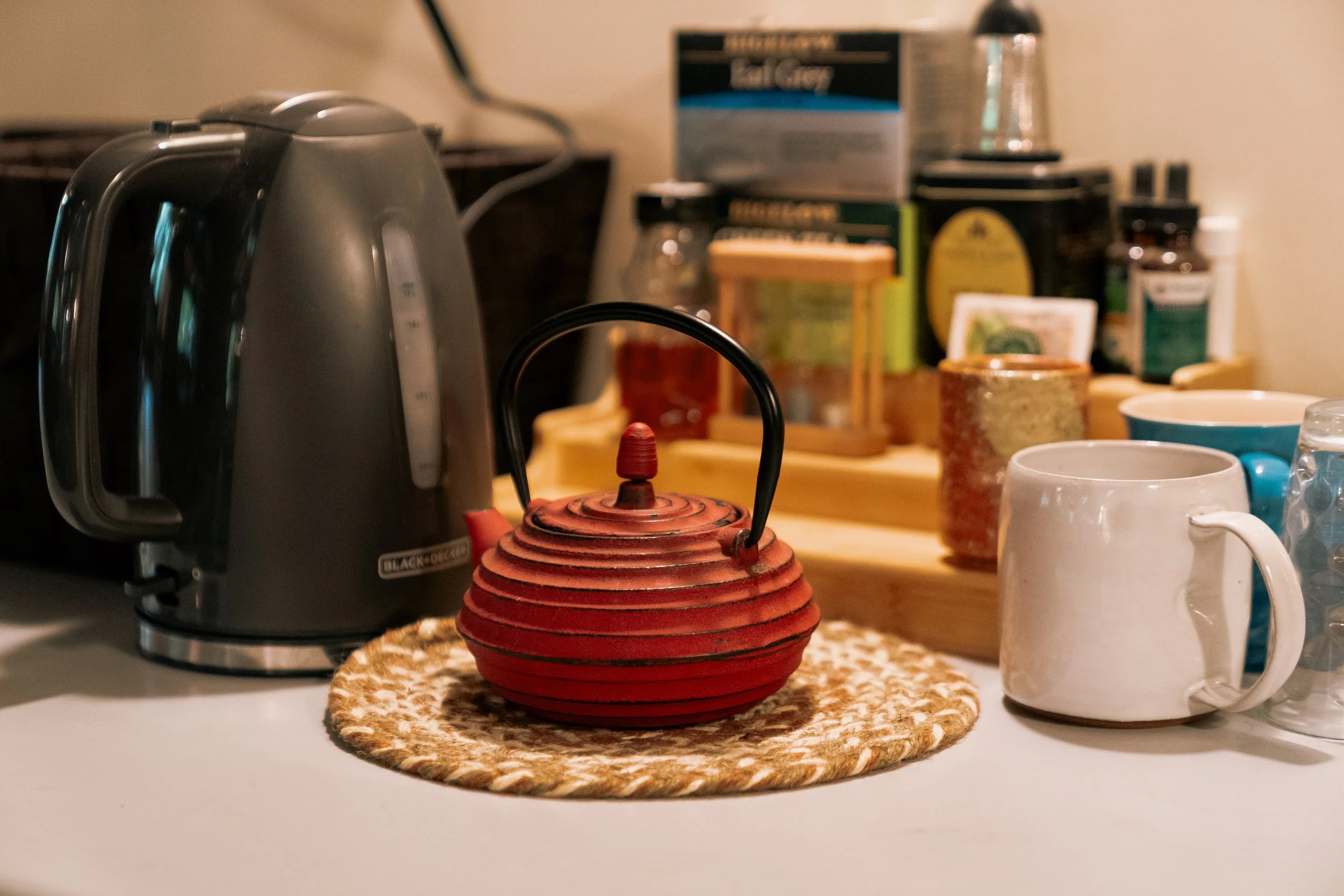 A kitchen countertop with a black electric kettle, a small red teapot, and various mugs and containers.