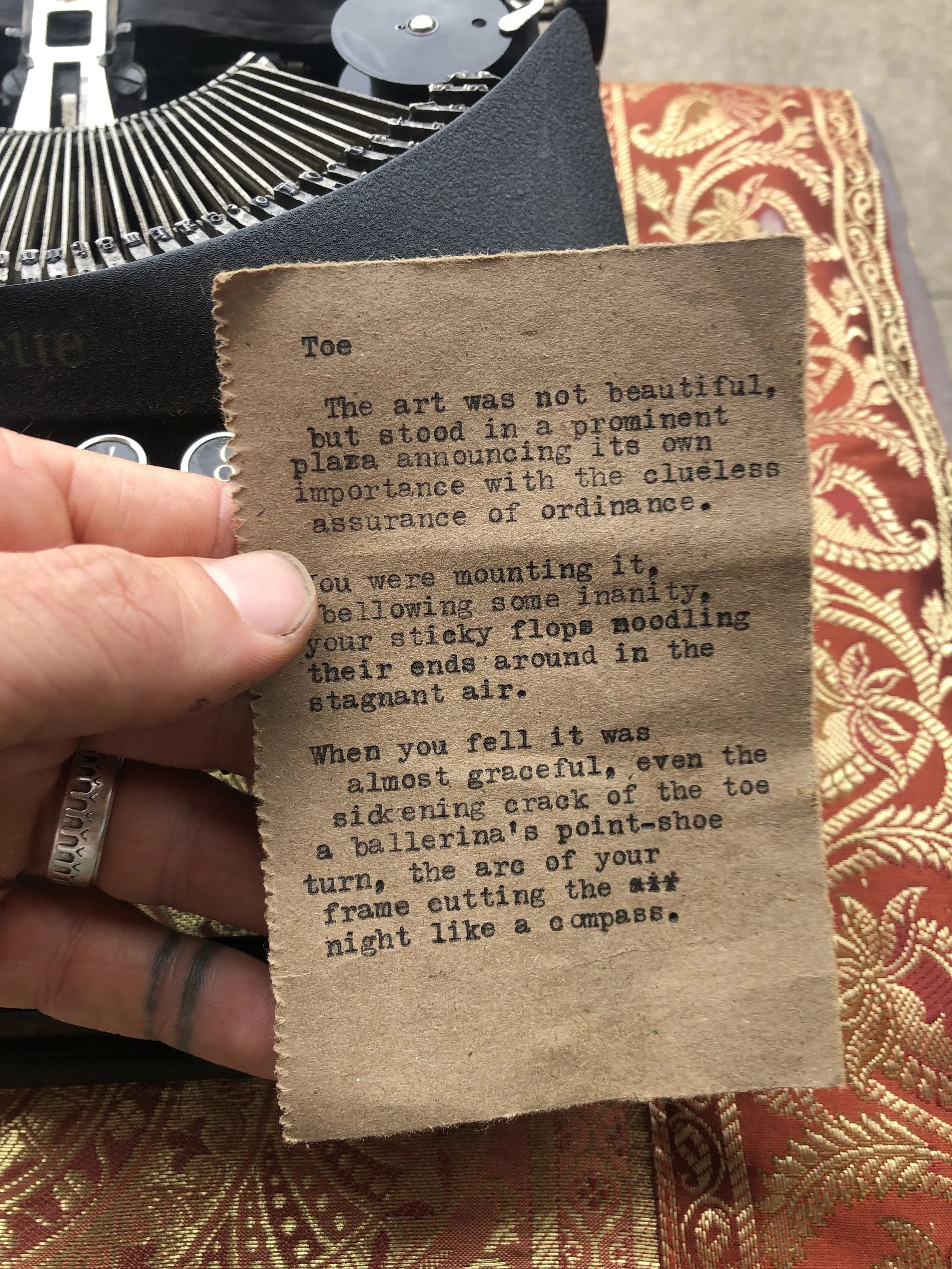 A person holding a small, torn piece of paper with typewritten text. In the background, a vintage typewriter and red and gold patterned fabric are visible.