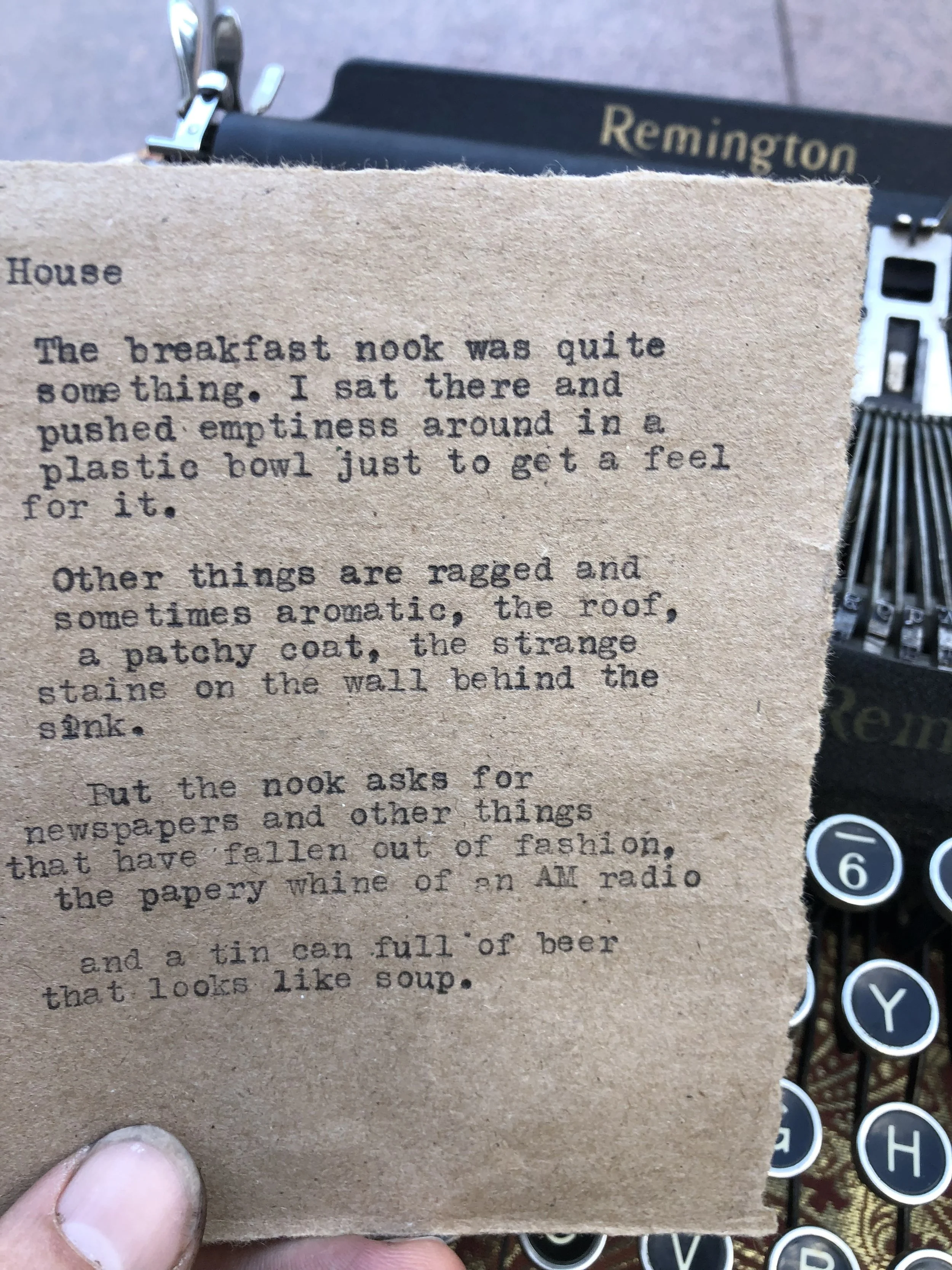 A close-up of a piece of paper with typewritten text held by a person's hand, with a Remington typewriter and what appears to be a wooden floor in the background.