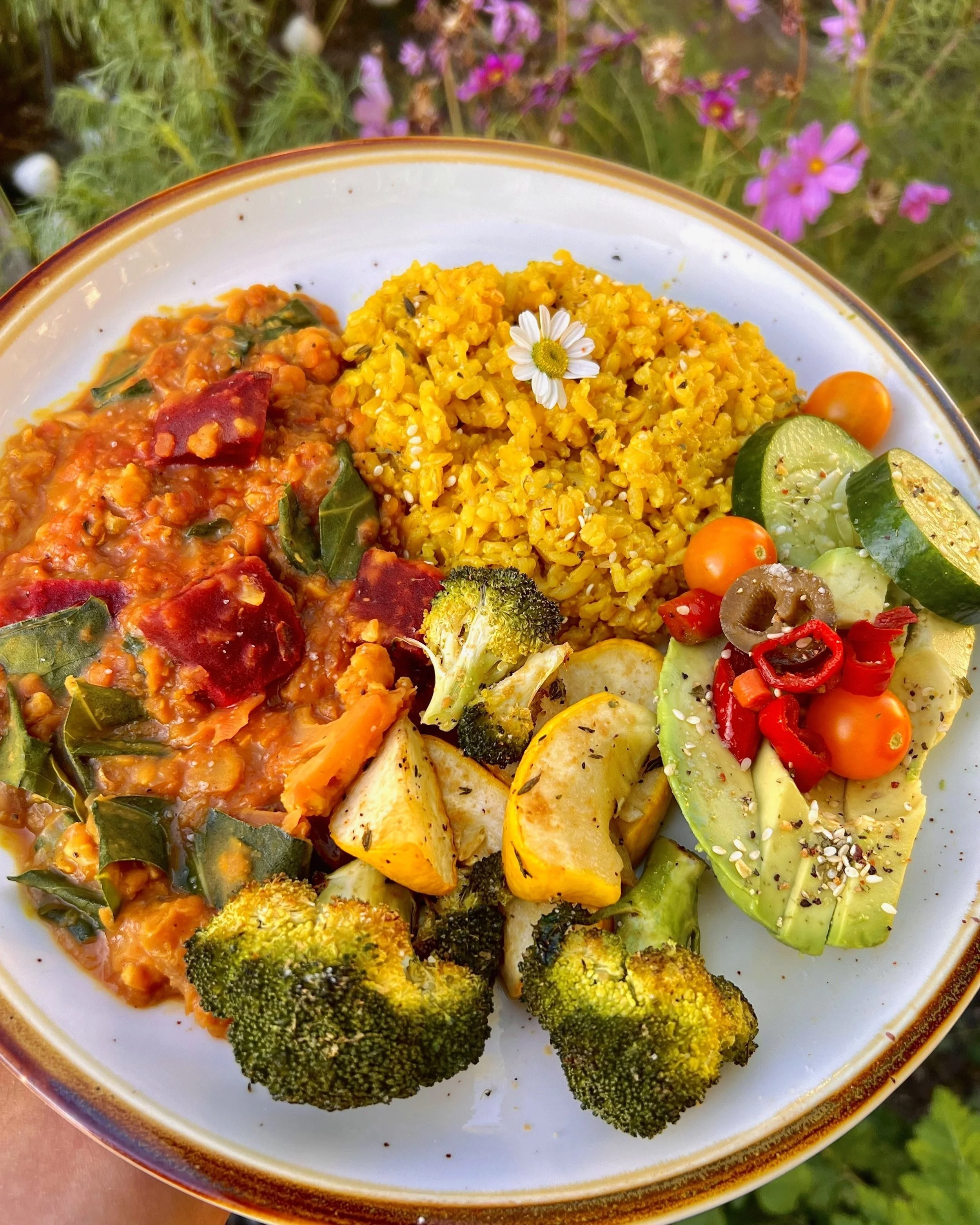 Plate of yellow rice, roasted broccoli, roasted mixed vegetables, and a salad with cherry tomatoes, cucumber, red pepper, and avocado slices, garnished with a small daisy flower.
