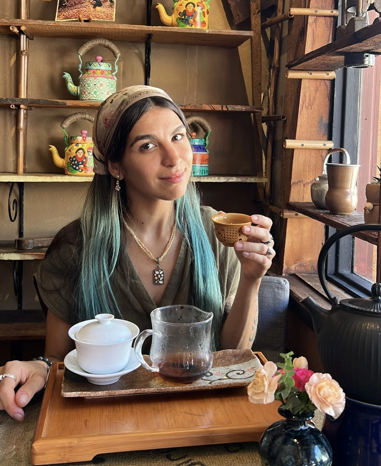 A woman with long hair dyed blue at the ends, wearing a headscarf, earrings, a pearl necklace, and a brown top, sitting at a wooden table with a tea set and a flower vase, holding a small tea cup, in a cozy room with wooden shelves decorated with colorful teapots.