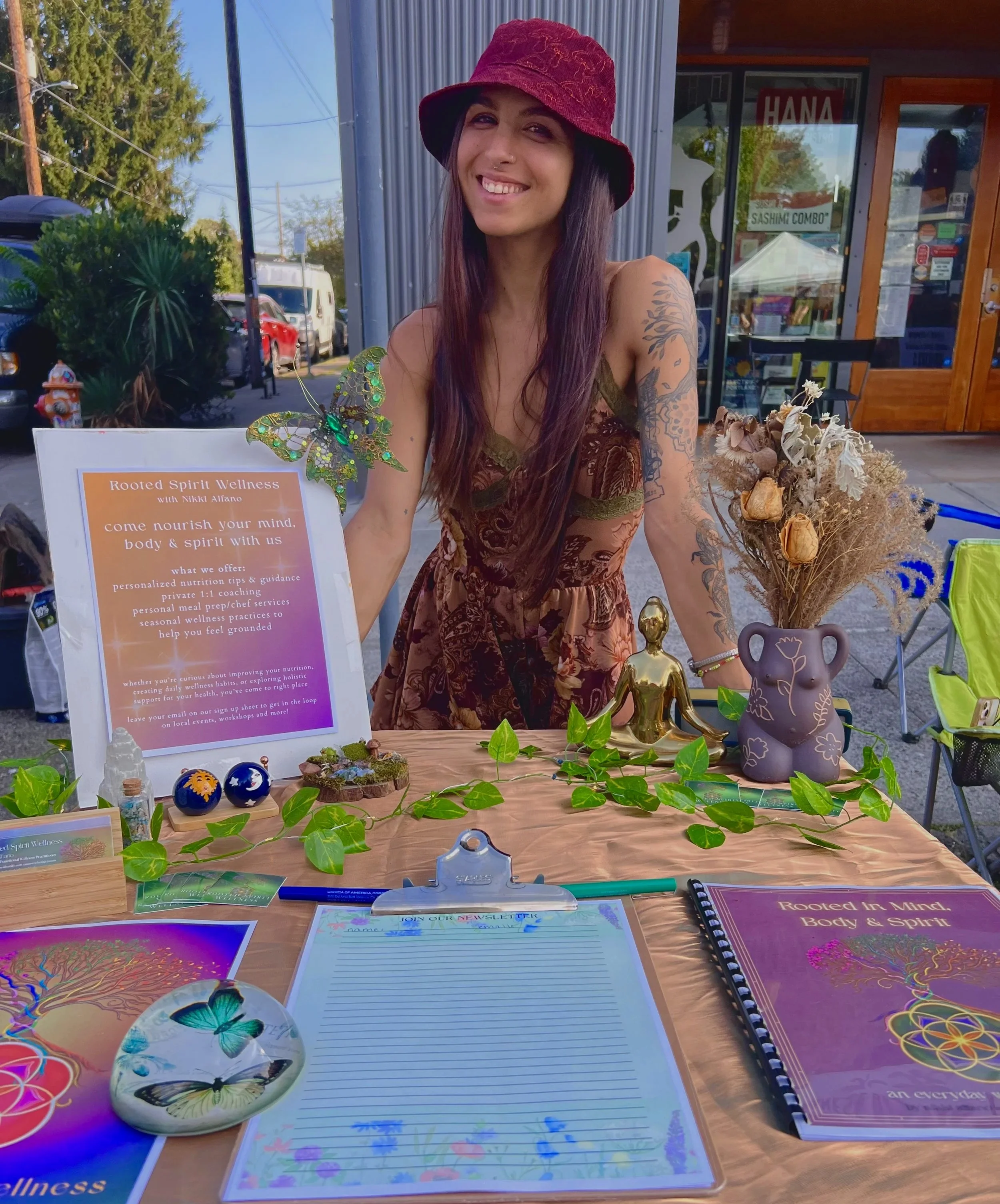 A woman with long dark hair, wearing a burgundy hat and a colorful dress, smiling at a wellness booth outside a storefront. The booth features a sign, a vase with dried flowers, a small golden statue, and colorful decorative items.
