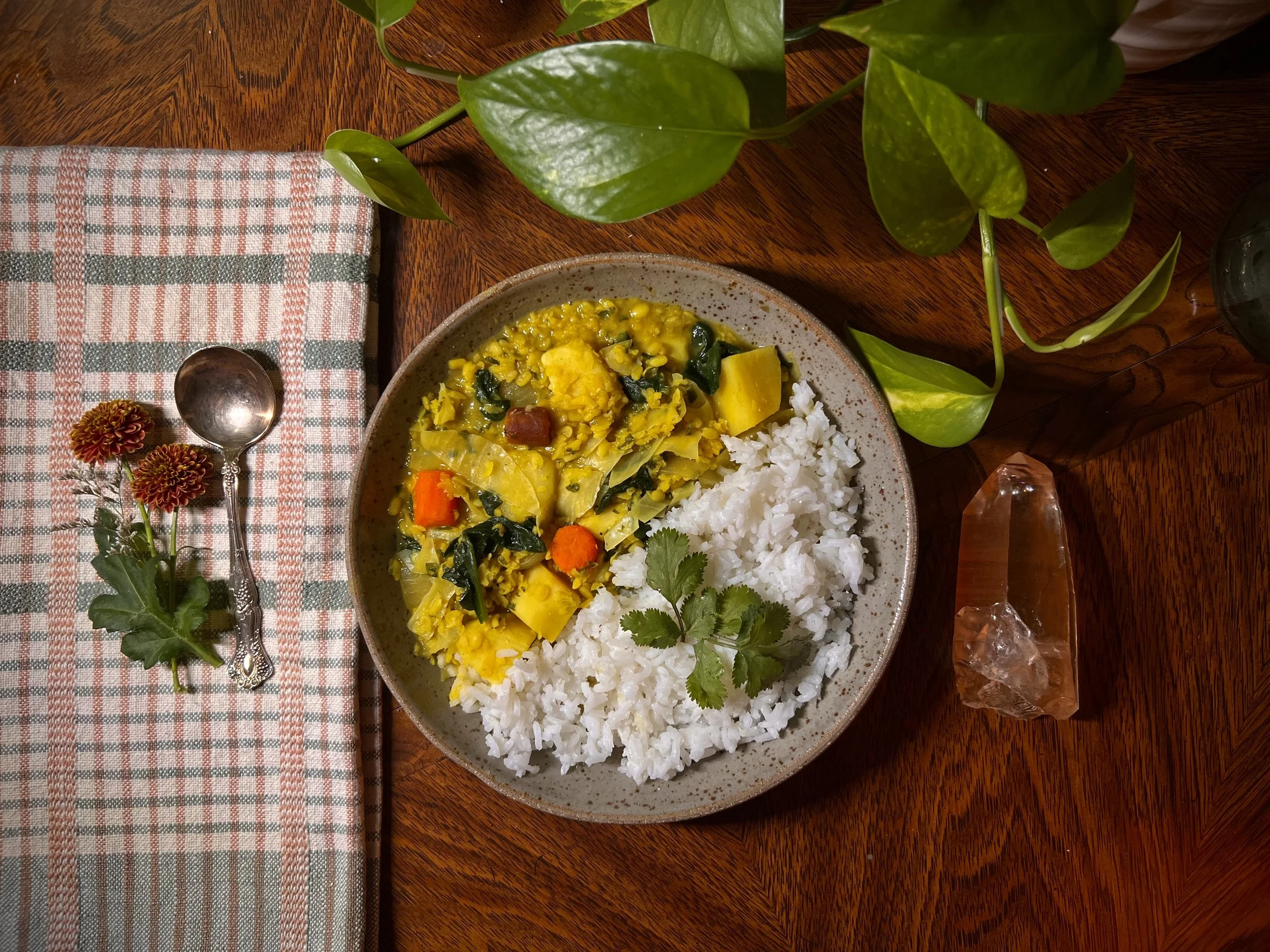 A bowl of rice and vegetable curry on a wooden table with a potted plant, a small spoon, a cloth napkin with flowers and a crystal on it.
