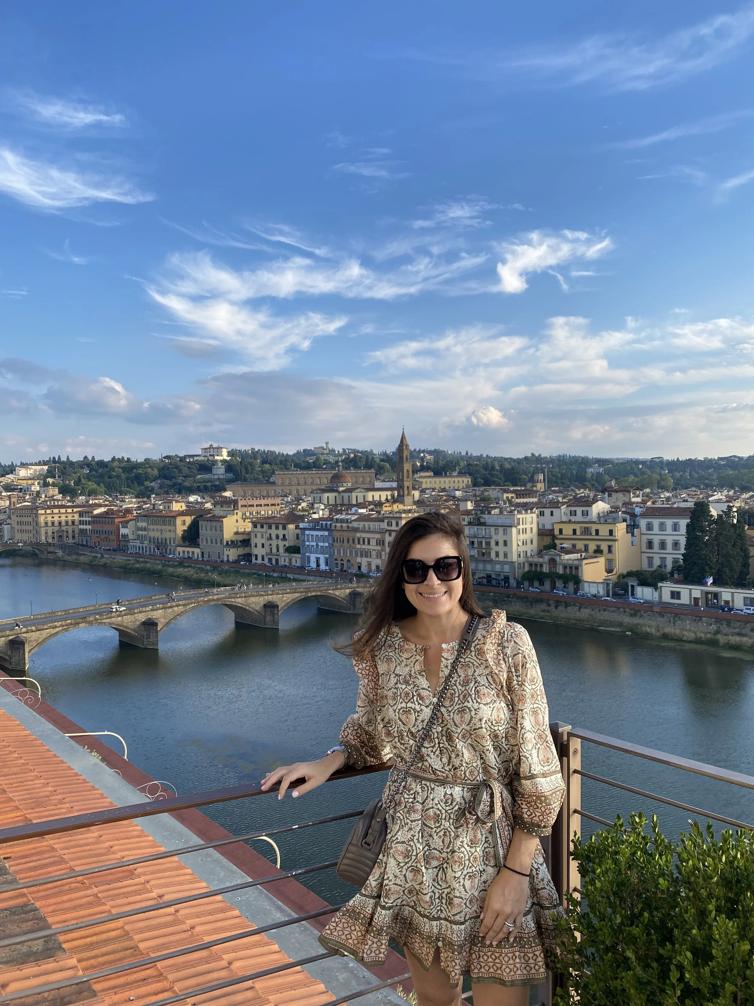 A woman with long brown hair and wearing sunglasses is smiling and posing near a river with a European cityscape in the background, including historic buildings, a bridge, and a church with a tall steeple.
