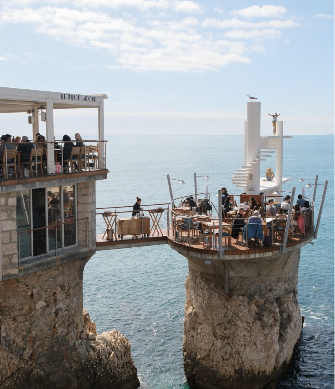 Two levels of a seaside restaurant with outdoor seating on rocky cliffs, overlooking the ocean, featuring modern design with white structures, umbrellas, and patrons dining amid a clear sky with scattered clouds.
