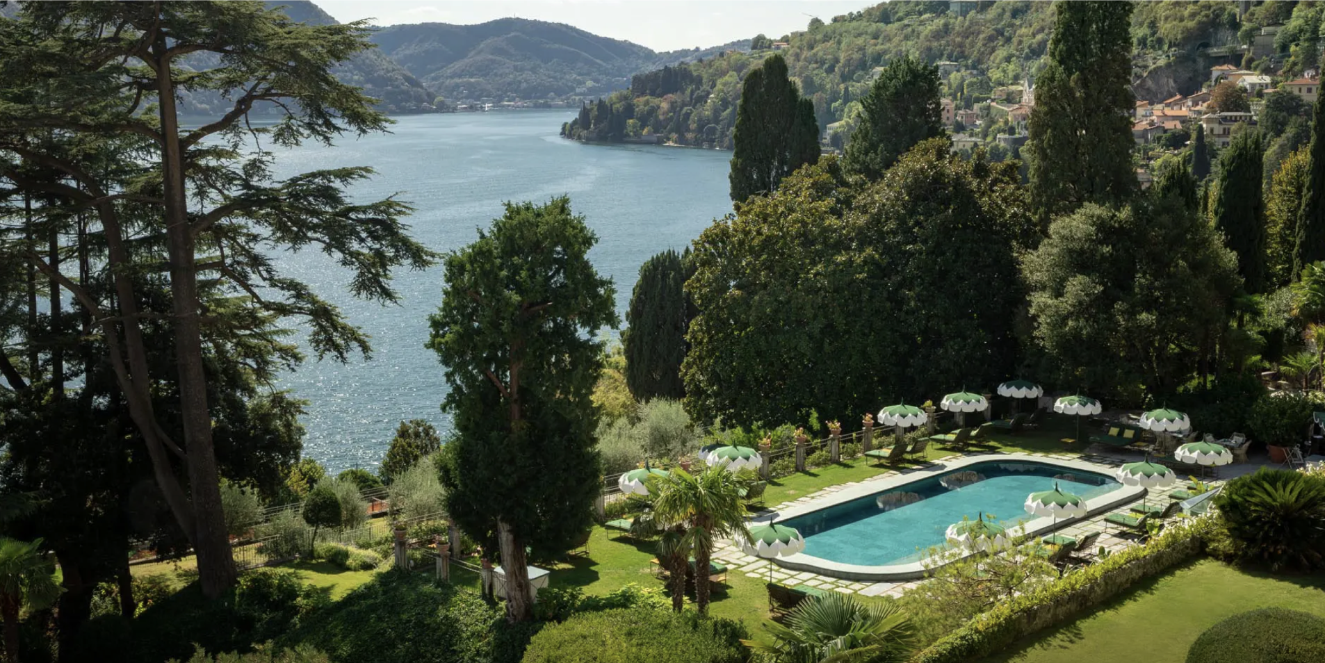 View of a scenic lakeside area with lush green trees, a swimming pool with white and green umbrellas, and a hillside town in the background.