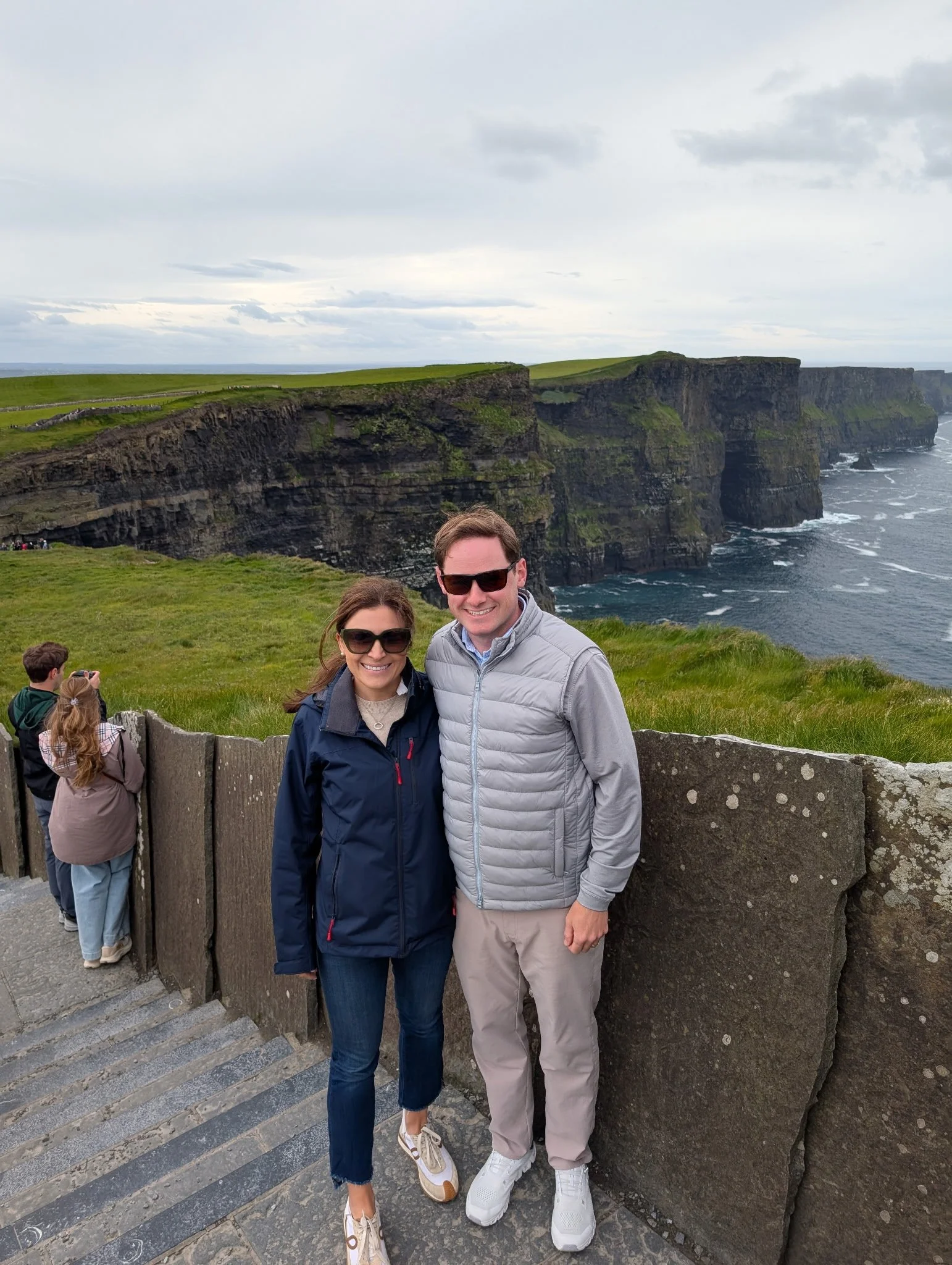A smiling couple stands on a stone staircase at a scenic cliffside overlook with green grass, rugged cliffs, and the ocean in the background. Other visitors are visible in the background.