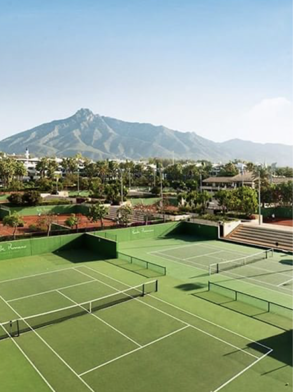 Tennis courts with multiple courts and nets, surrounded by green fences, with residential buildings and trees in the background, and a mountain range in the distance under a clear blue sky.