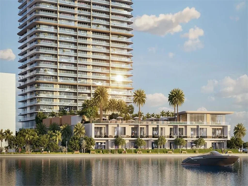 Modern high-rise building with glass balconies and palm trees in front, next to a waterway with a yacht, under a partly cloudy sky.