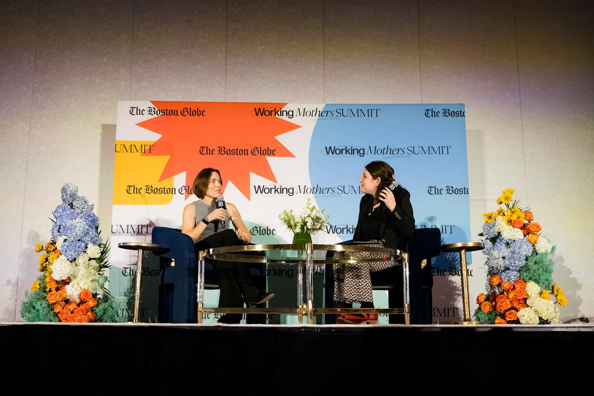 Two people speaking at a summit while sitting on a stage with floral arrangements on either side of them