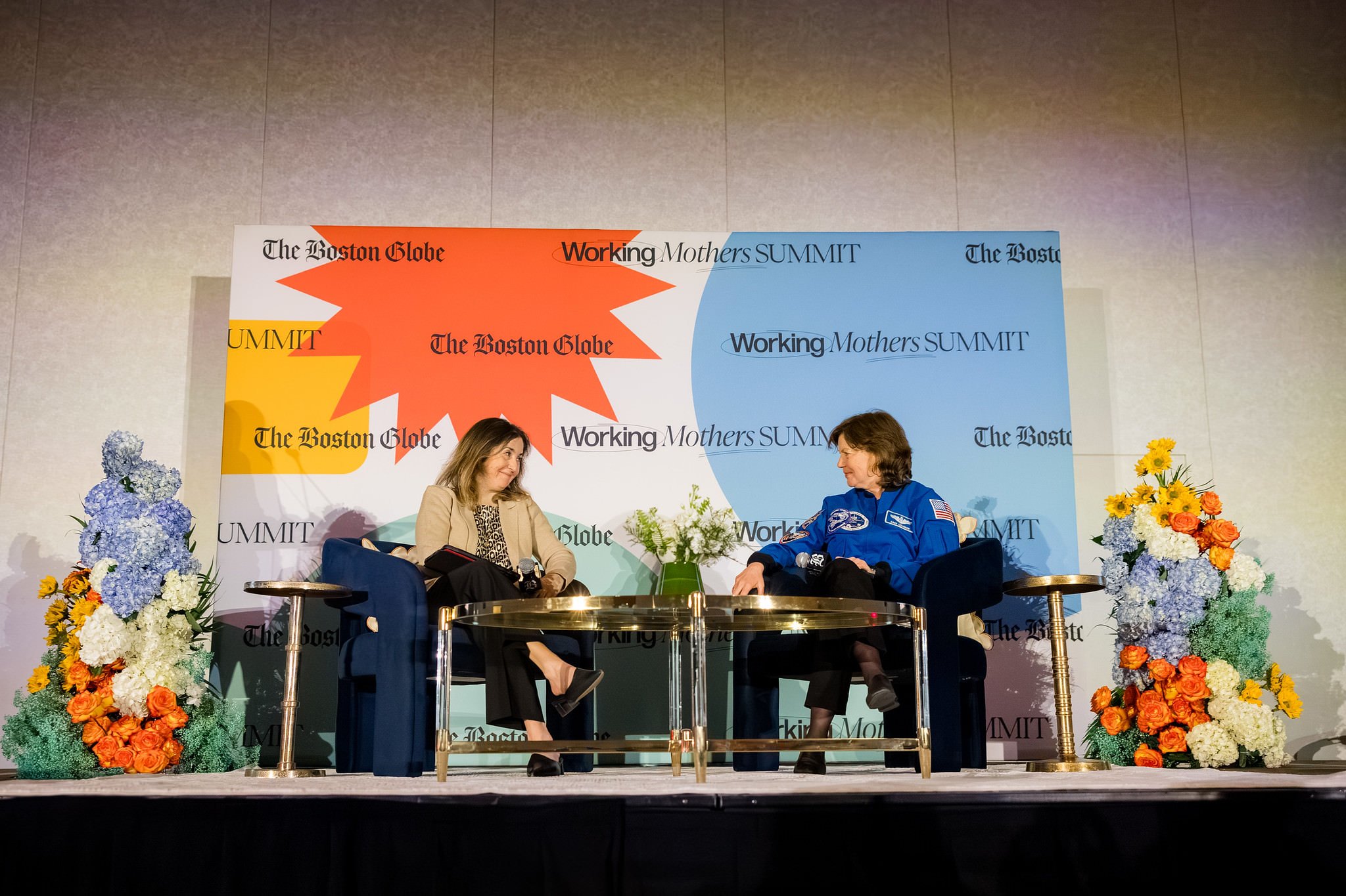  Two people speaking on a stage with floral arrangements on either side of them  