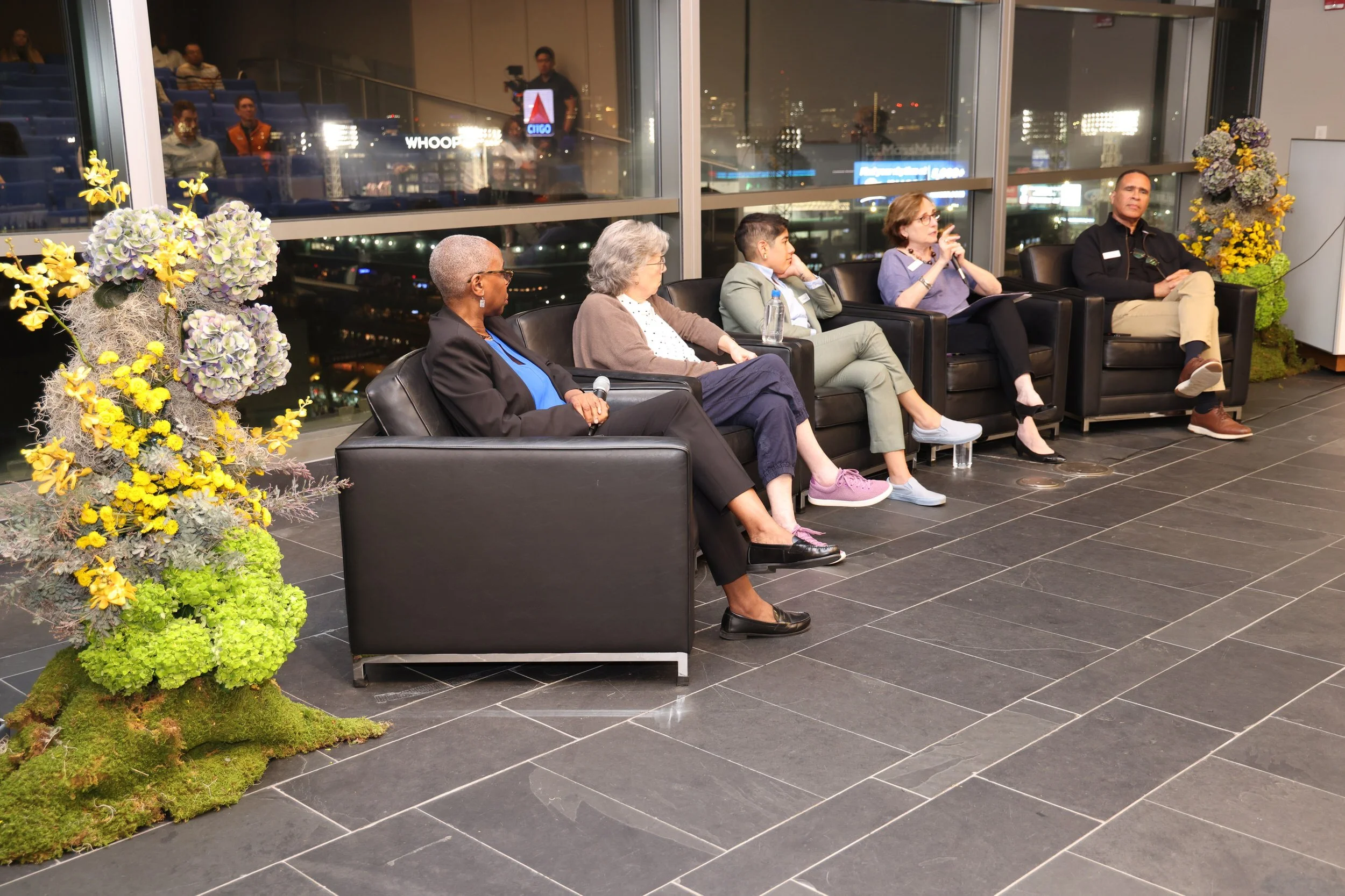  A panel of speakers sitting in front of a window with floral arrangements on either side of them  