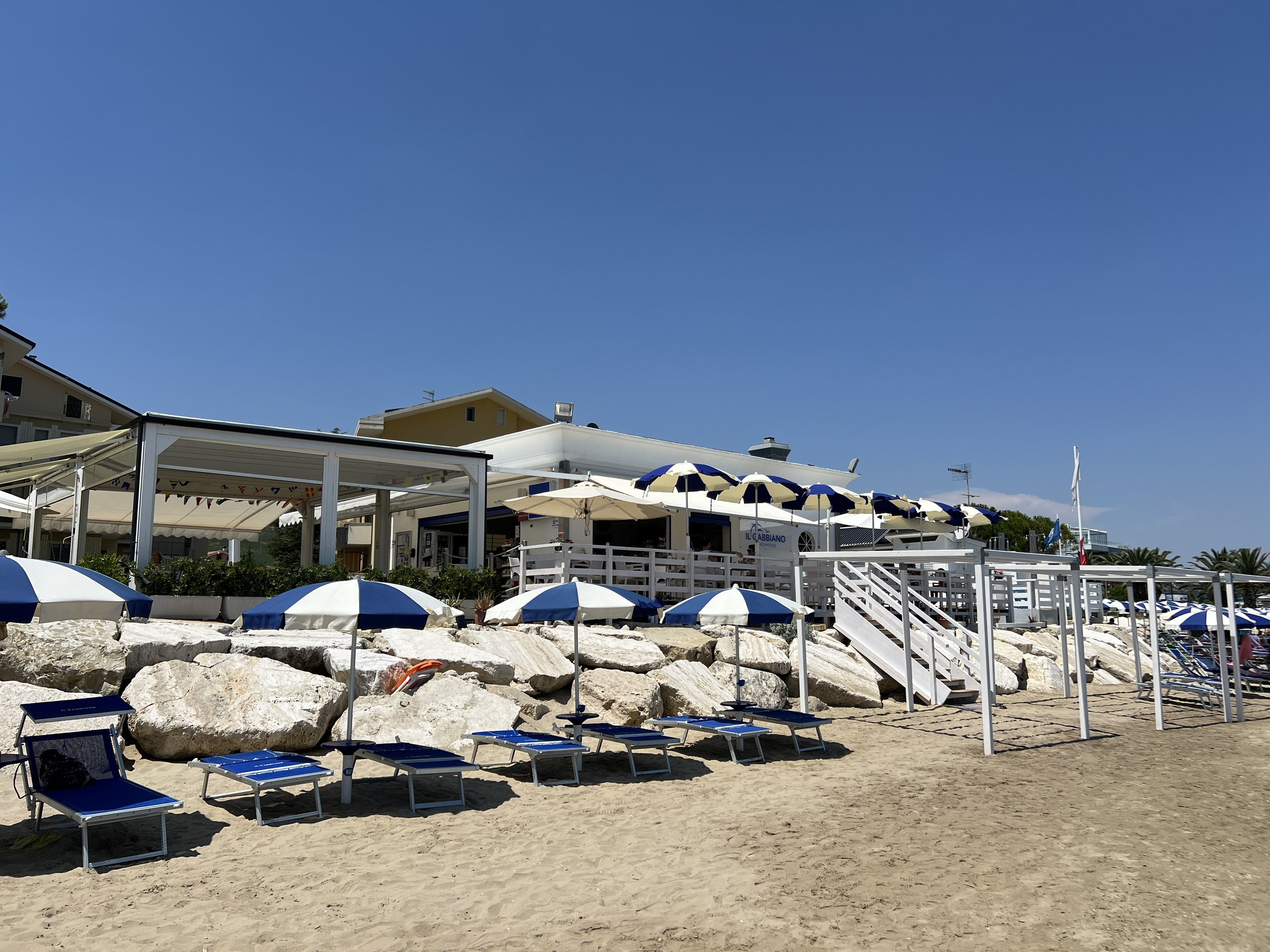 Spiaggia con ombrelloni blu e bianchi e lettini di fronte a un ristorante con terrazza.
