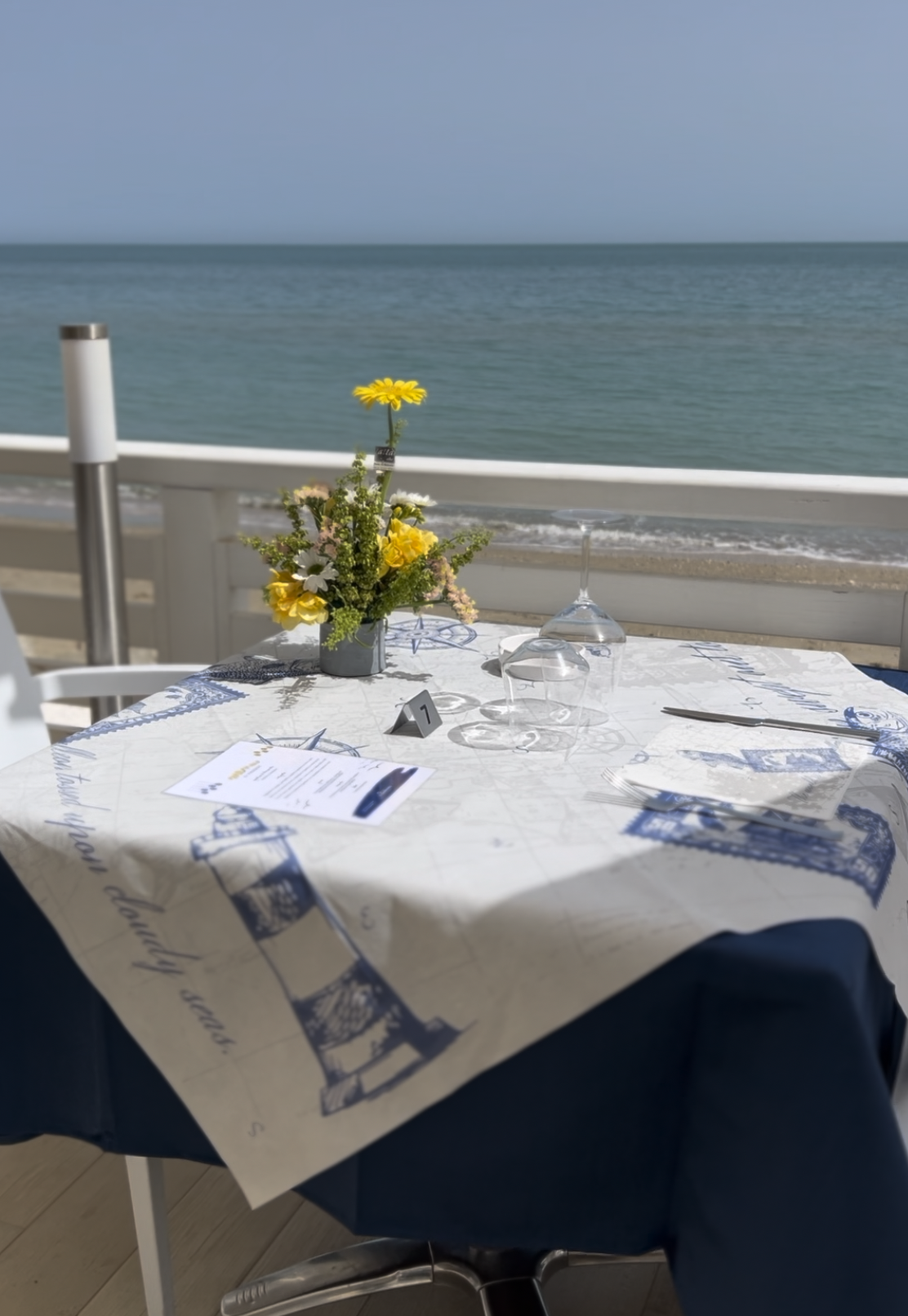 Tavola apparecchiata con un mazzo di fiori gialli, bicchieri di vino capovolti, un menu e posate, con vista sul mare