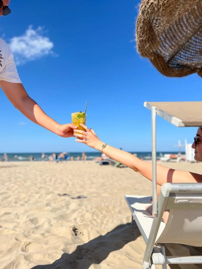 Una persona seduta su una sedia a sdraio in spiaggia che riceve un cocktail da un'altra persona. Sullo sfondo, il mare e il cielo blu con alcune nuvole.