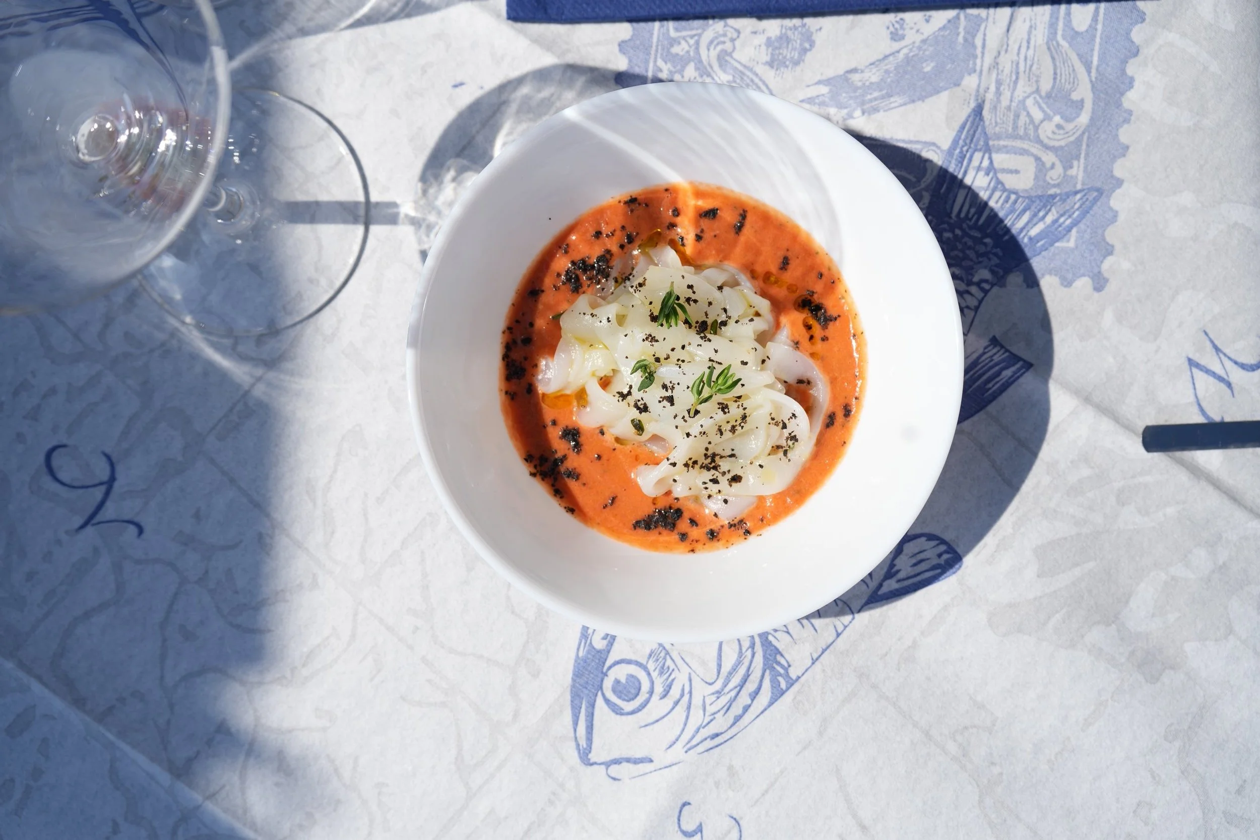 Piatti di pasta con salsa rossa, guarniti con erbe e pepe, posati su un tovagliolo con disegni di pesci, con bicchieri di acqua e vino accanto.