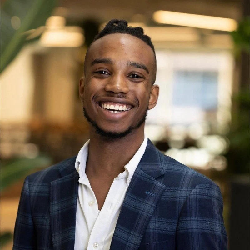 A smiling young man with short dreadlocks, wearing a blue plaid blazer and a white collared shirt, standing indoors with warm lighting and blurred background.