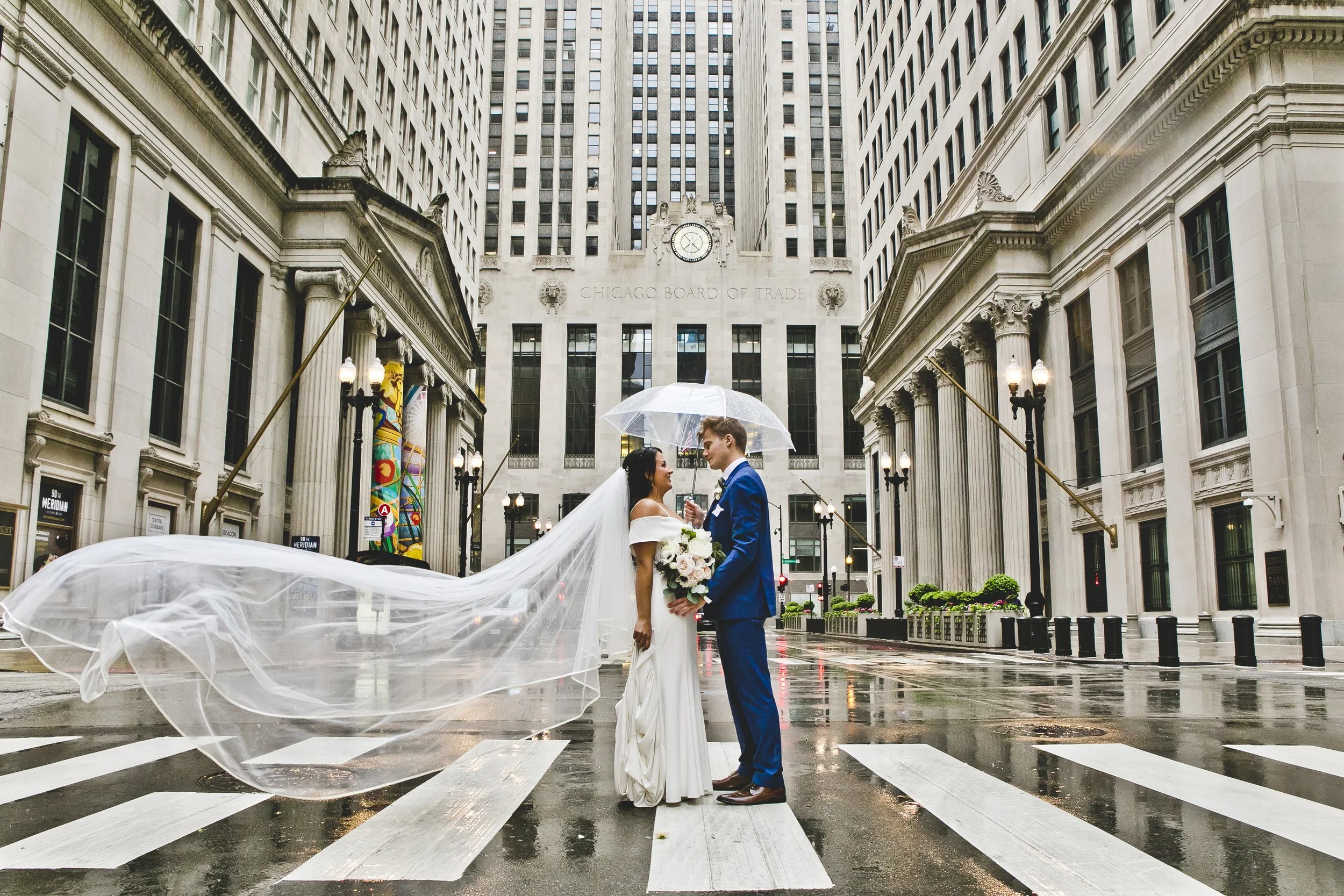 Bride in a white wedding gown standing outdoors, holding her dress as she poses for a photo near trees and city buildings.