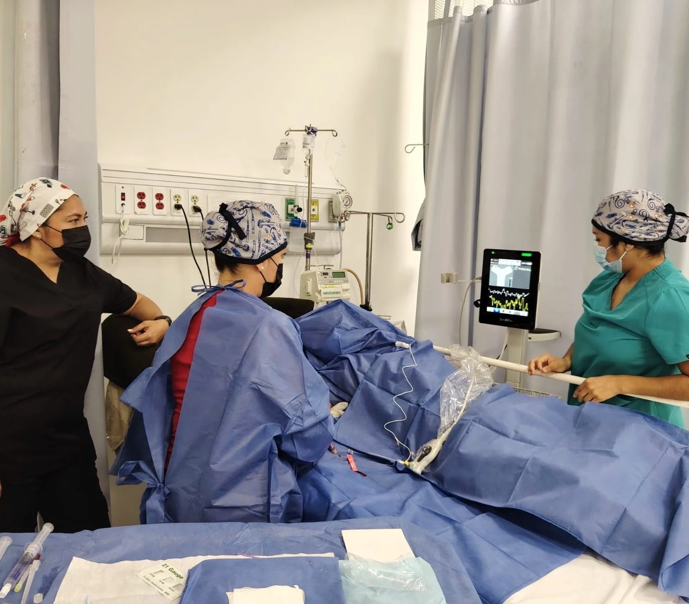 Personal médico en un hospital atendiendo a un paciente en la cama, con monitores y equipo médico a su alrededor. Todos usan mascarillas y gorros quirúrgicos. ACÉRCATE Asociación para certificación y capacitación Técnica Catéter PICC