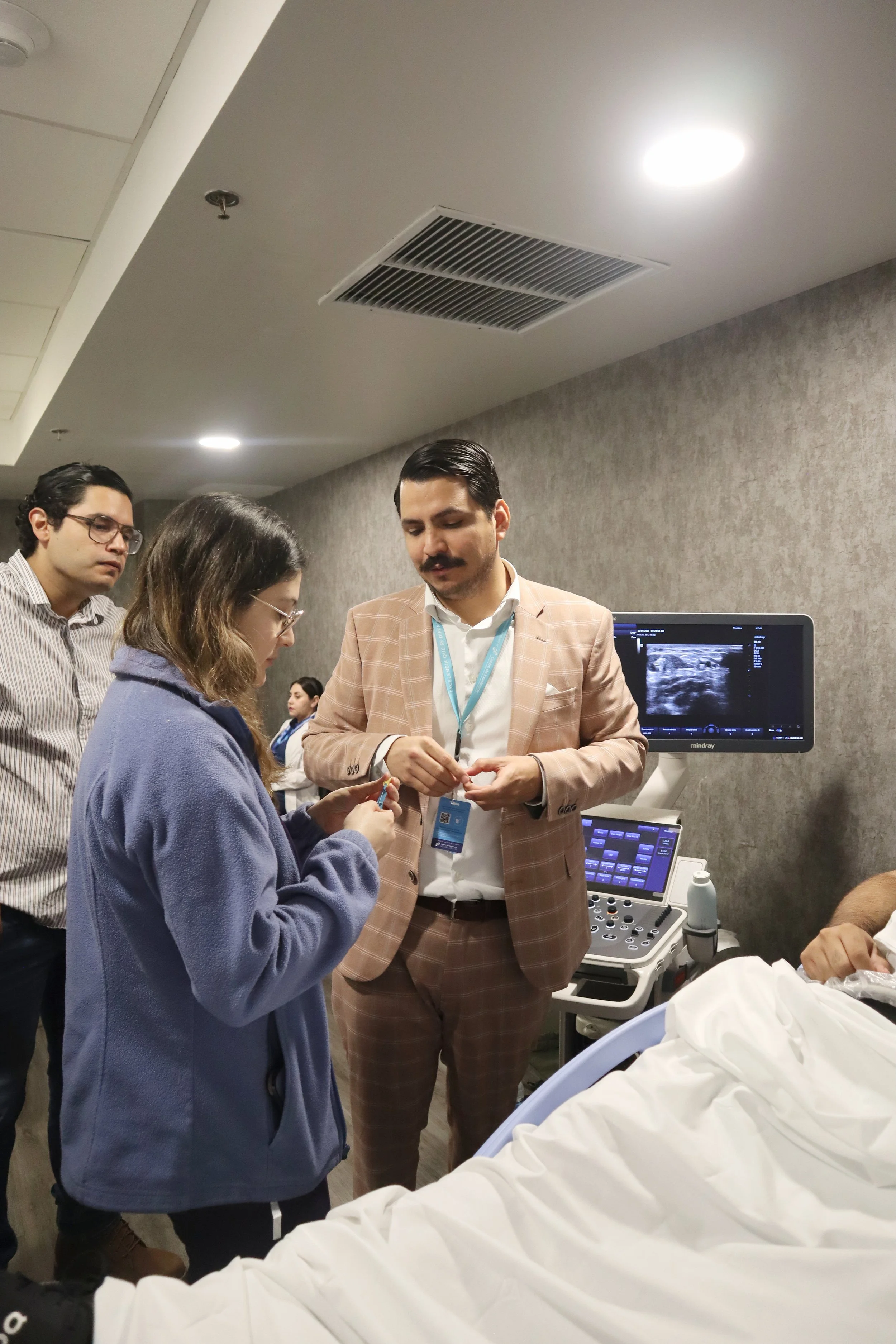 Médicos y enfermero conversando a la cabecera de un paciente en una sala de hospital, con monitor de ultrasonido en la pared. ACÉRCATE Asociación para certificación y capacitación Técnica Catéter PICC