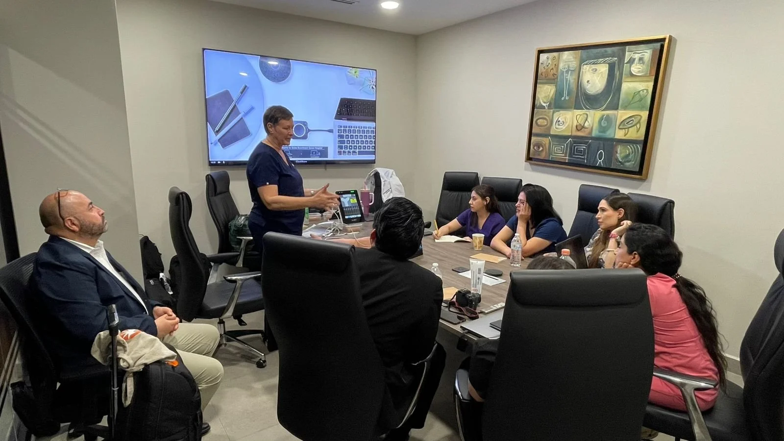 Reunión en una sala de conferencias con personas sentadas en una mesa, una mujer de pie frente a una pantalla grande mostrando una presentación, y varios objetos en la mesa como laptops, botellas de agua y cuadernos. ACÉRCATE Catéter PICC
