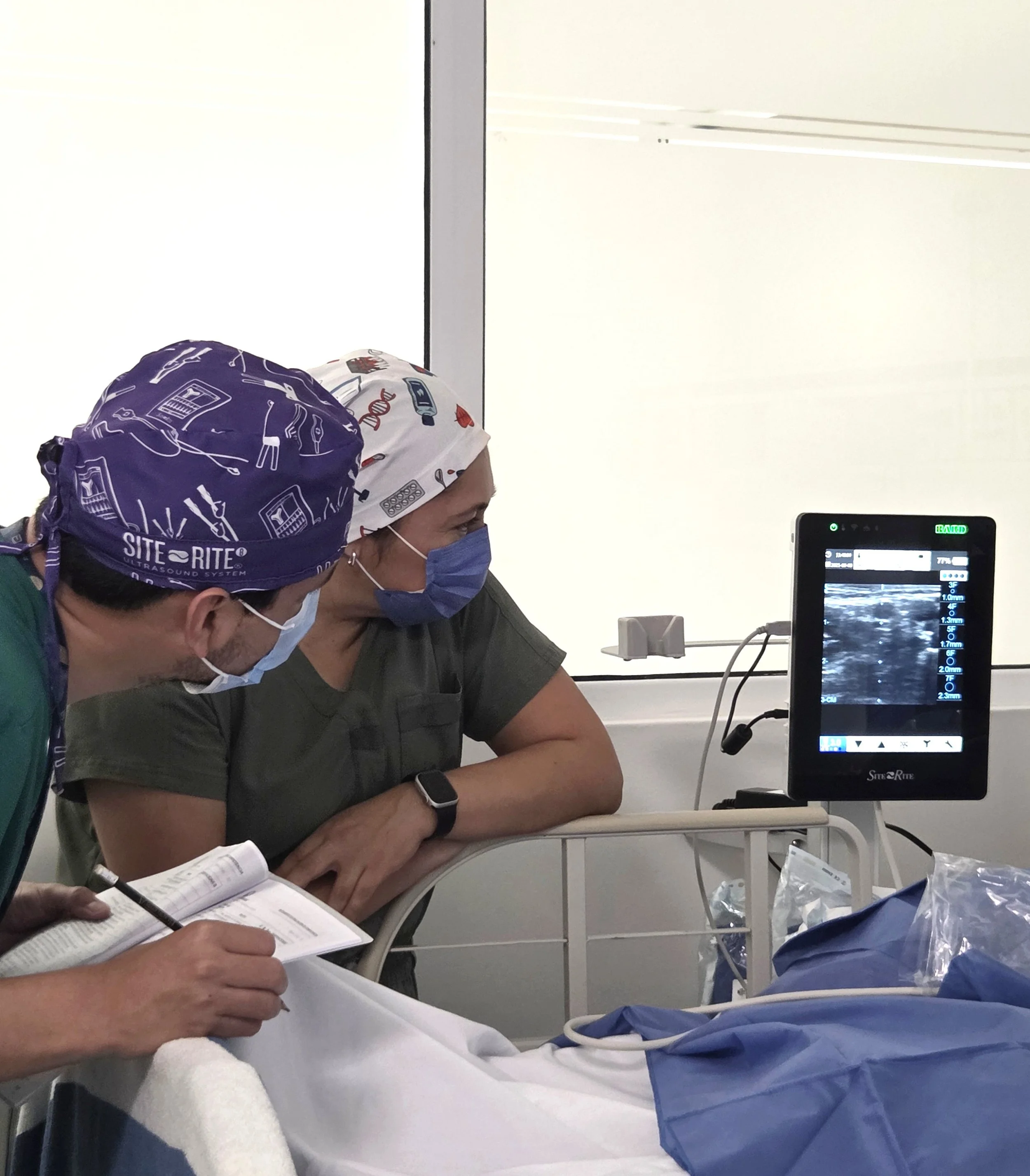 Médicos en un hospital revisando una ecografía en una pantalla digital, ambos usan mascarillas y gorros médicos. ACÉRCATE Catéter PICC