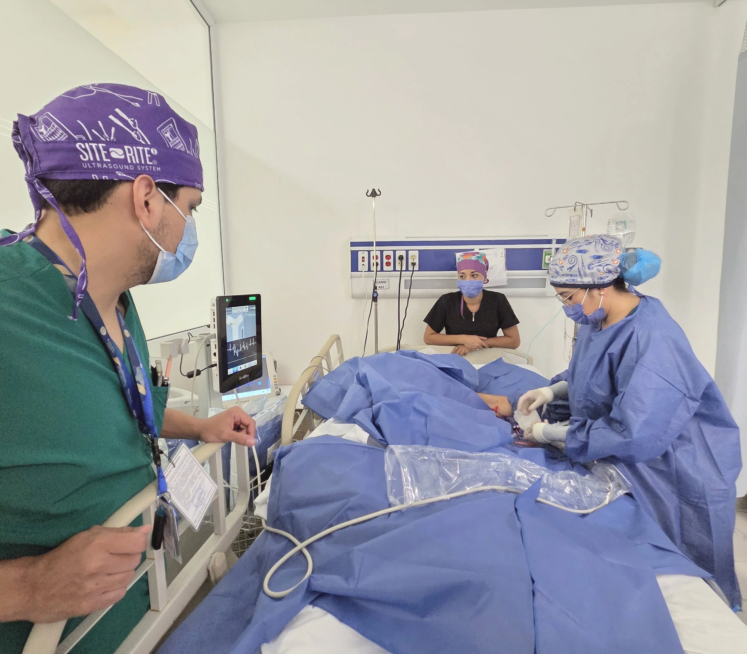 Equipo médico en una sala de hospital atendiendo a un paciente en cirugía, con monitor y equipo médico en el fondo. ACÉRCATE Catéter PICC