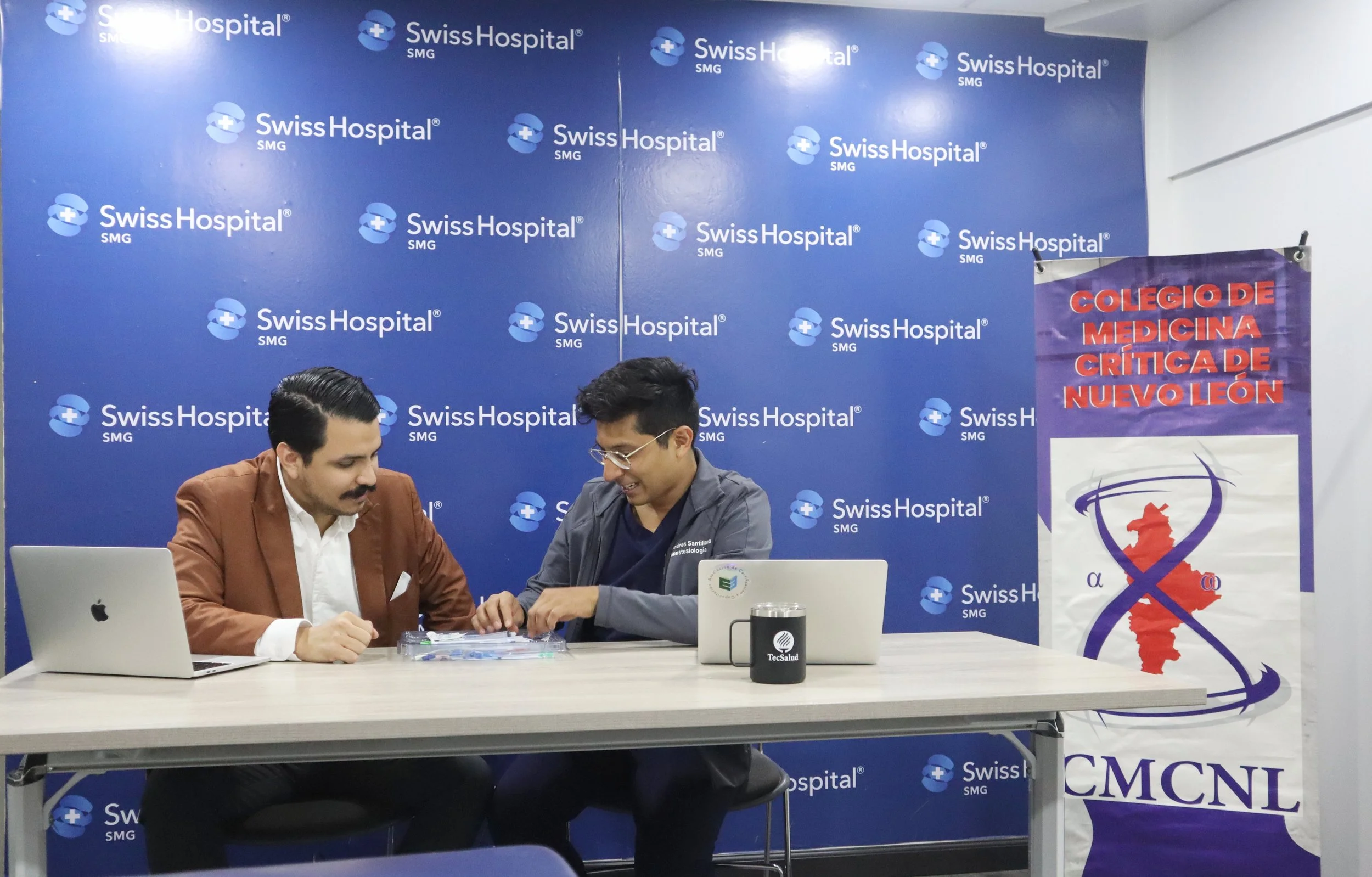Dos hombres en una clínica o hospital, uno con bata y el otro con saco, conversando sobre un asunto médico, frente a una mesa con computadoras y documentos. Detrás, un fondo azul con el logo de Swiss Hospital. ACÉRCATE para PICC