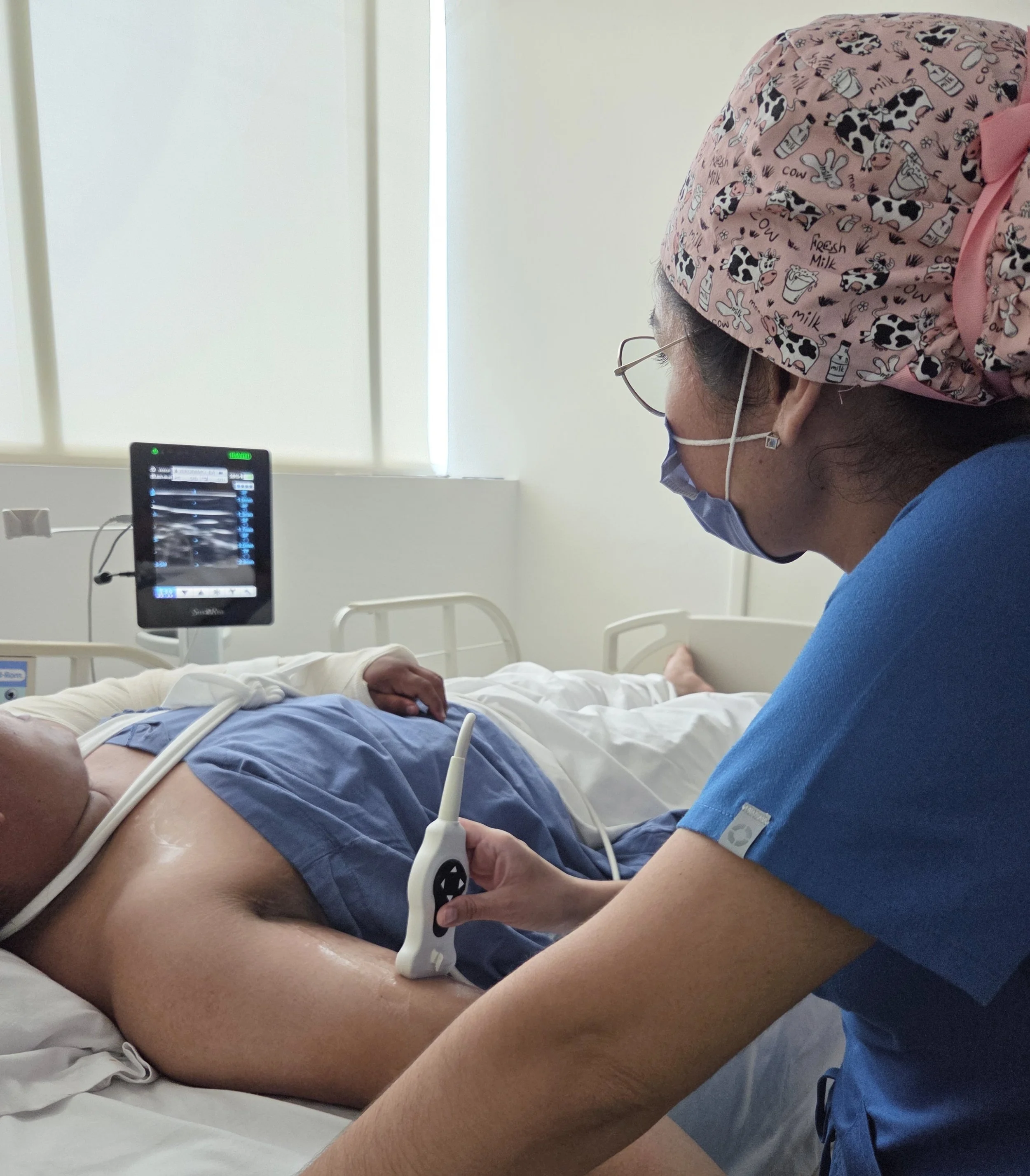Enfermera realizando una ecografía en un paciente en una cama de hospital, con monitor y ventanas de fondo. ACÉRCATE Catéter PICC
