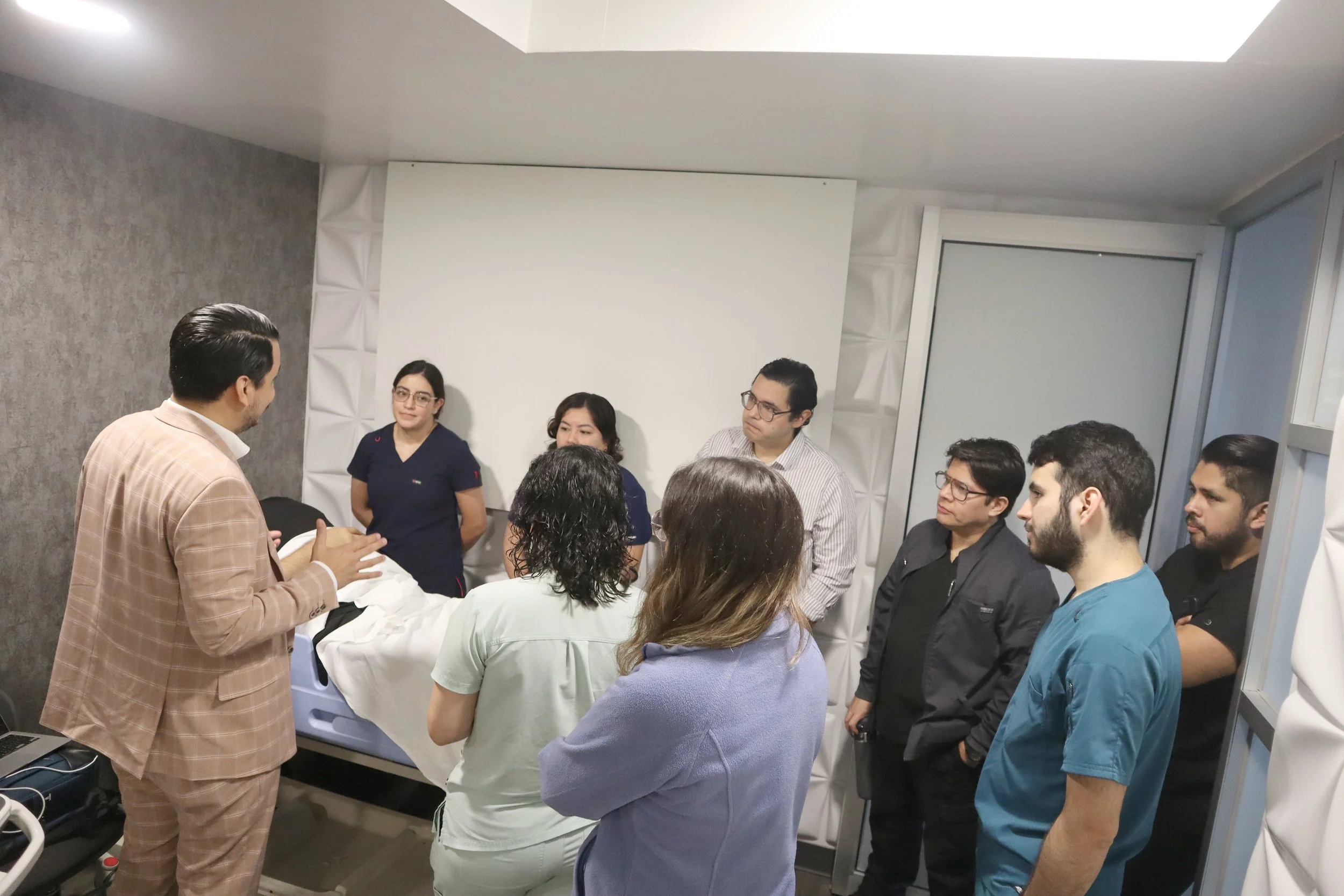 Grupo de personas en una sala de capacitación médica, atendiendo a un instructor que conversa con ellos, con una camilla en primer plano y paredes acolchonadas. ACÉRCATE Asociación para certificación y capacitación Técnica Catéter PICC