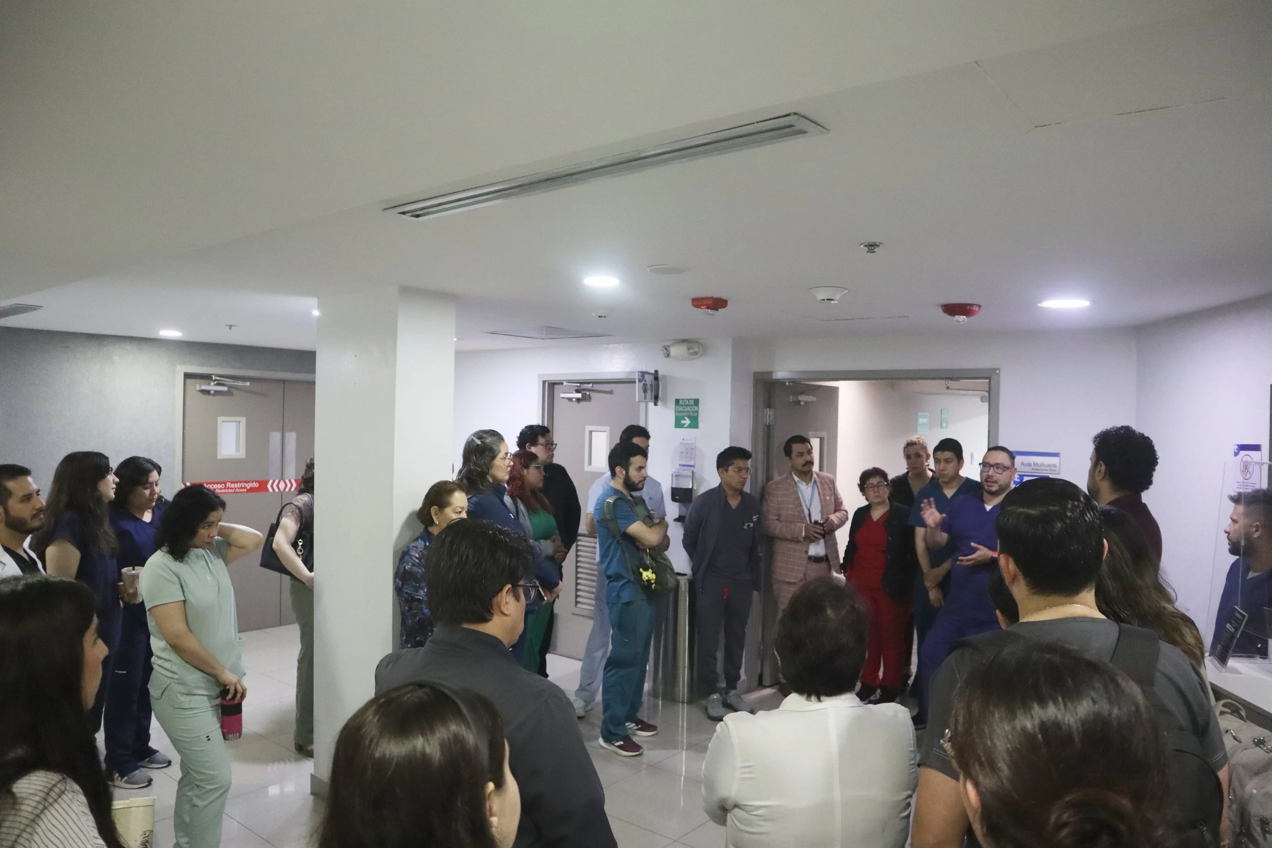 Un grupo de personas en un pasillo de un edificio, participando en una charla o reunión, algunas con gafas y otras con mochilas, escuchando a una persona que habla y está de pie. ACÉRCATE Asociación para certificación y capacitación Técnica Catéter P