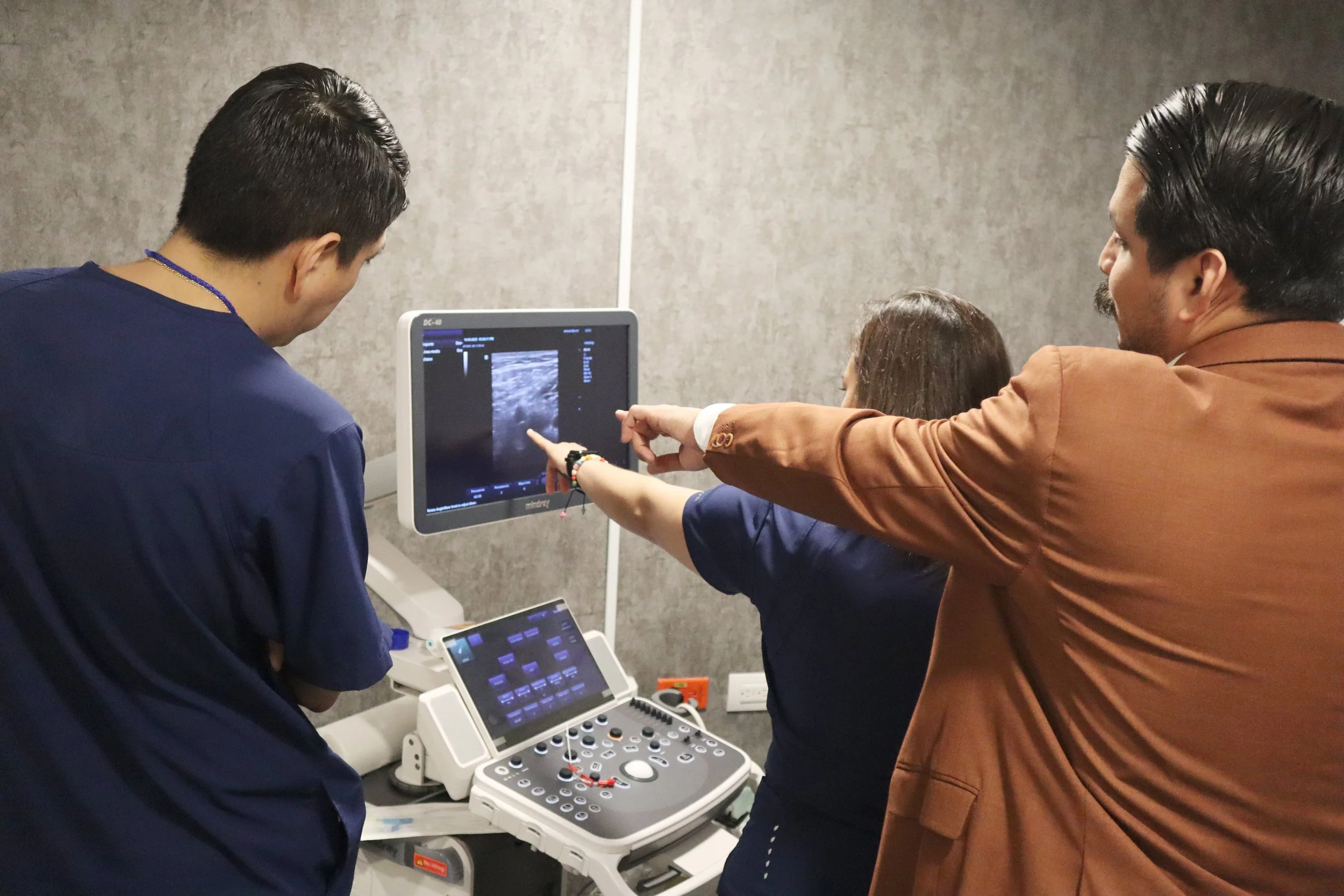 Tres personas en una consulta médica observando una pantalla de ultrasonido, una persona señala la imagen en la pantalla. ACÉRCATE Asociación para certificación y capacitación Técnica Catéter PICC