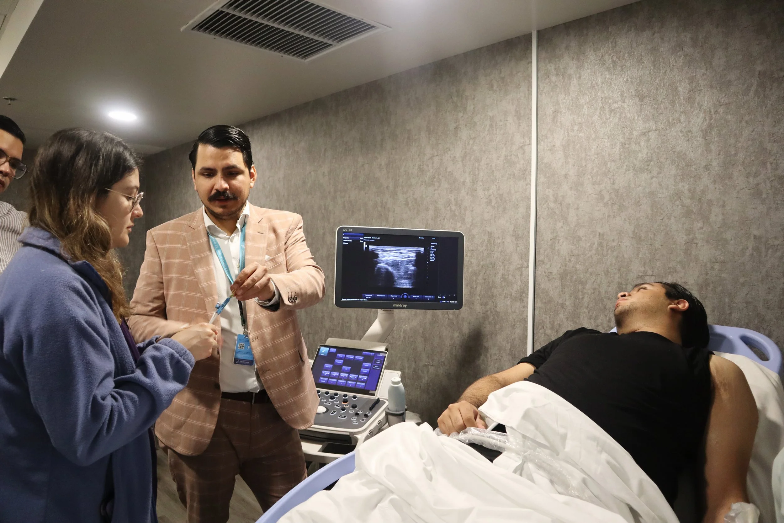 Un paciente en una cama de hospital con la cabeza levantada, rodeado de médicos y un estudiante en un ambiente clínico, con un monitor de ultrasonido. ACÉRCATE Asociación para certificación y capacitación Técnica Catéter PICC